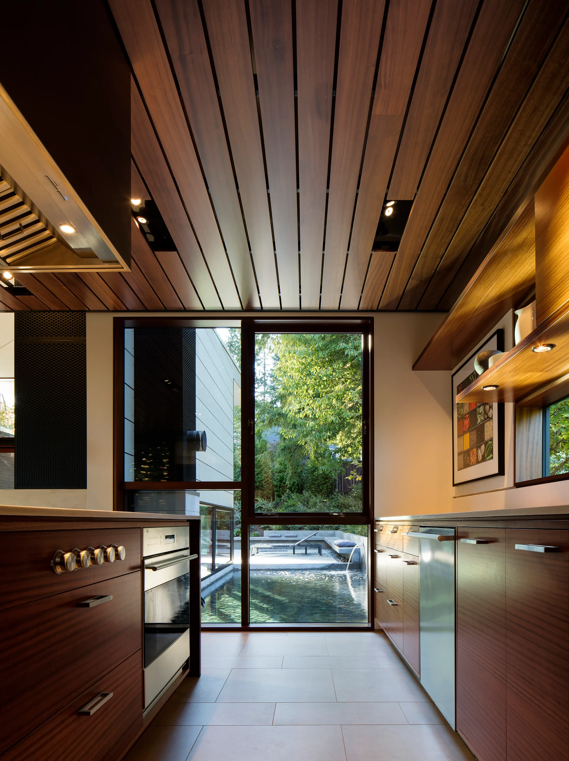A galley kitchen featuring dark wood cabinetry, a linear wood plank ceiling, and a large window looking out onto a backyard garden pond.