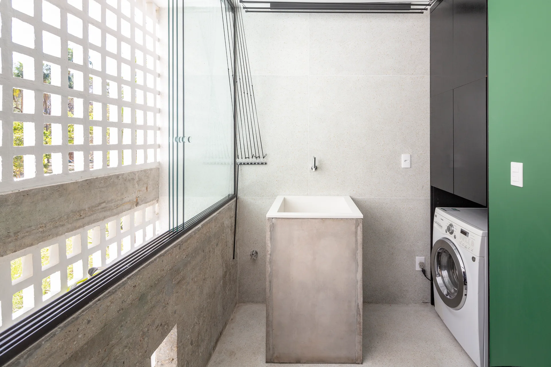 A minimalist utility room features a central raw concrete sink, a front-load washing machine, and a white breeze block screen wall.
