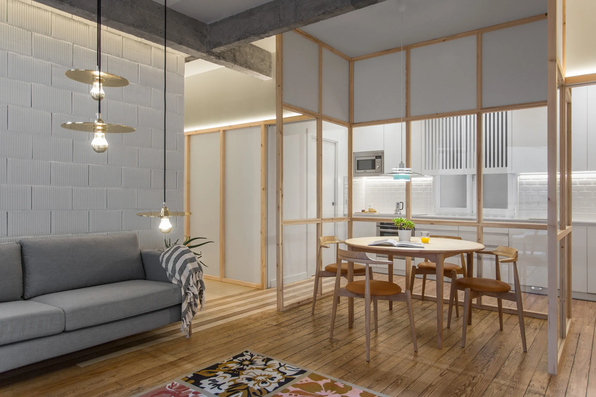 A modern living room features a gray sofa, brass pendant lights, and a wood-framed glass partition enclosing a dining table and kitchen.