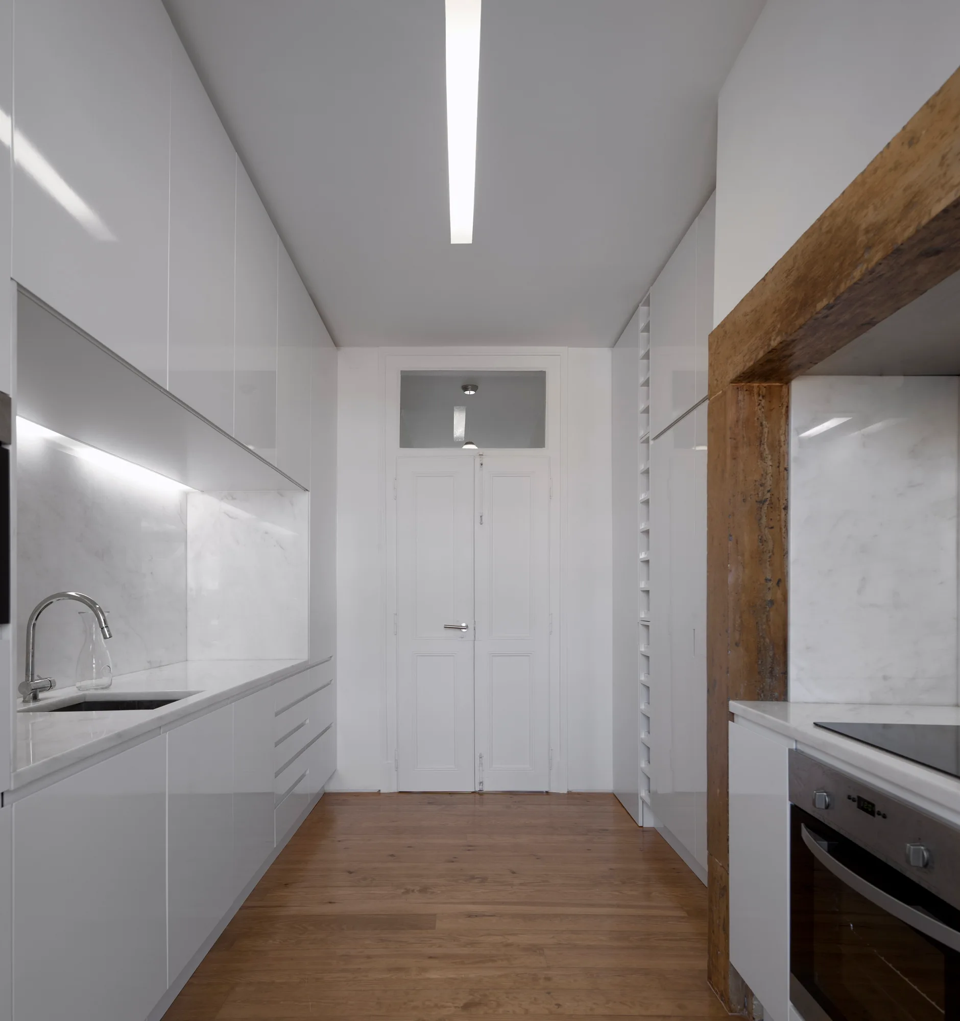 A modern galley kitchen featuring high-gloss white cabinets, a marble backsplash, and a rustic wooden beam framing an oven.