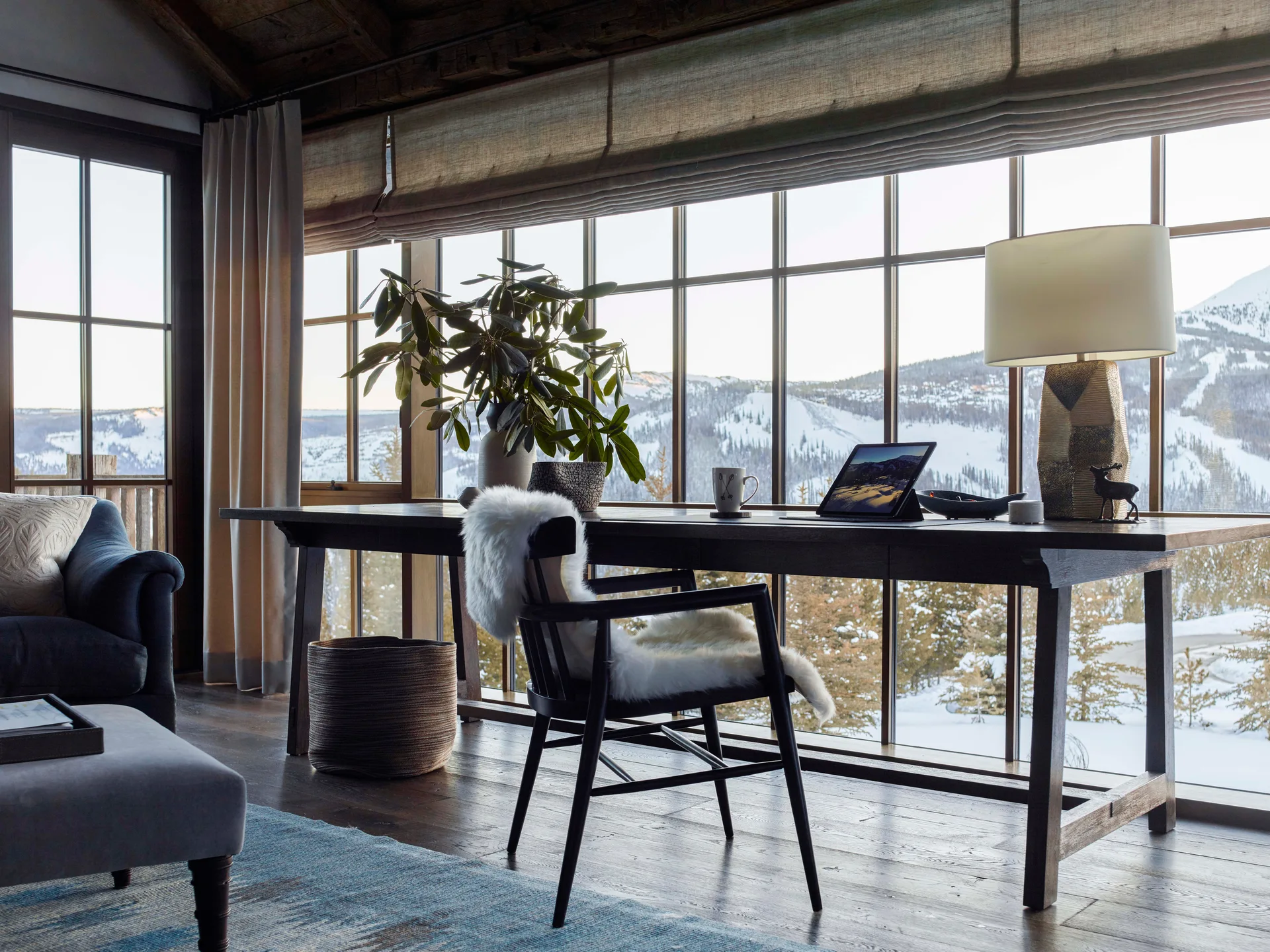 A rustic home office featuring a dark wood writing desk and a black wooden chair with a white sheepskin throw in front of a grid window wall.