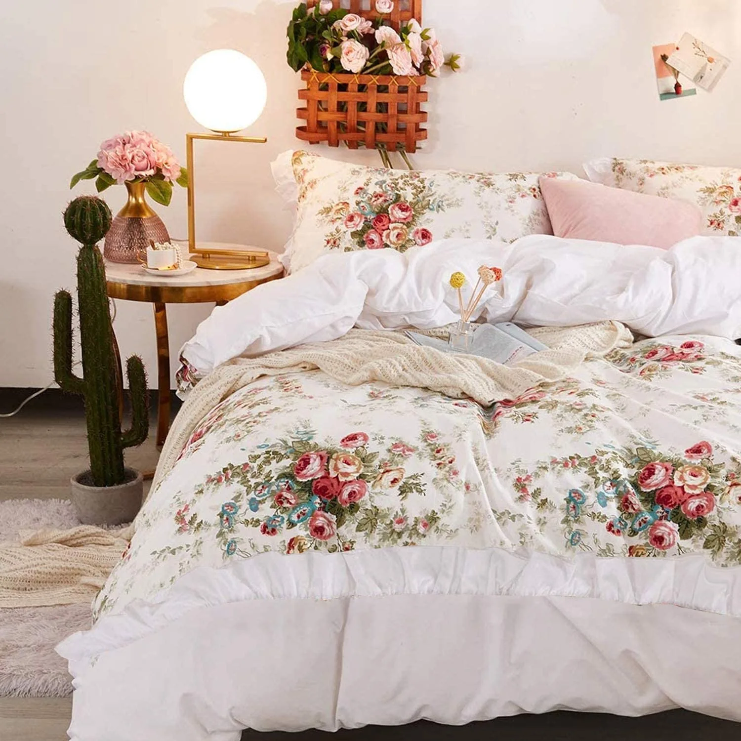 A bedroom bed styled with rose-patterned floral bedding and a white duvet next to a gold bedside table and a tall potted cactus.