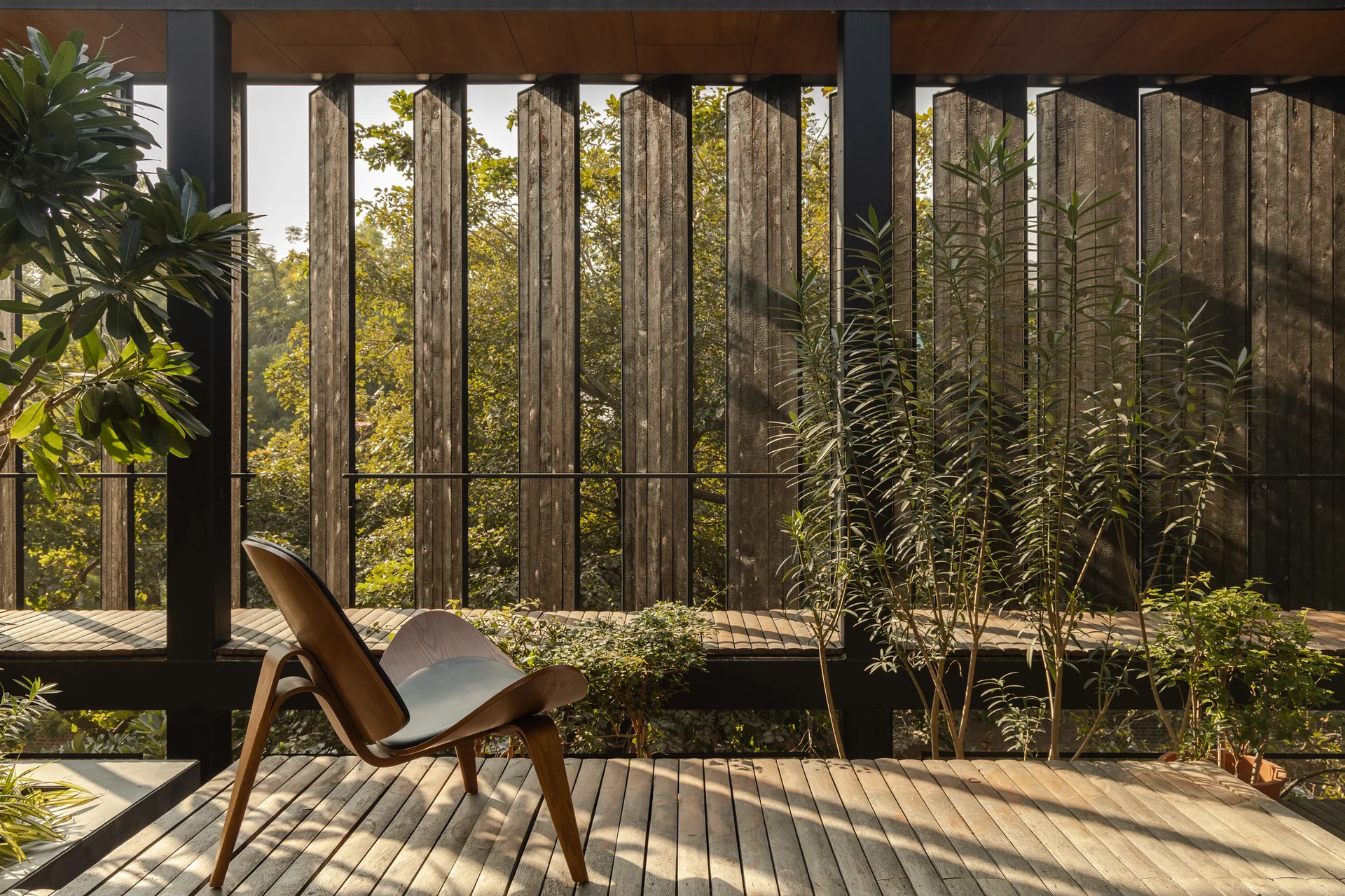 A curved wood lounge chair sits on a wooden deck next to potted plants, with a vertical wood slat privacy screen filtering sunlight.