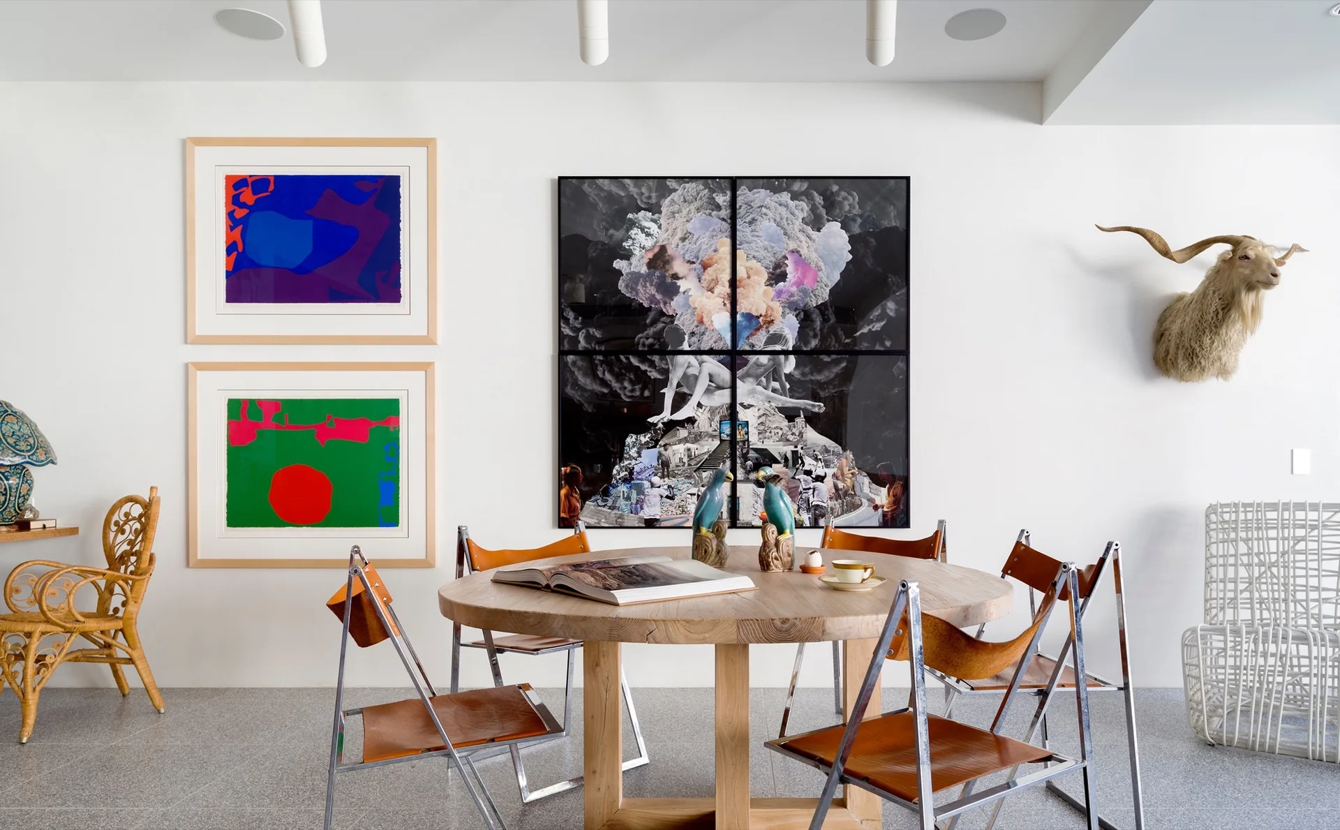 A round wooden dining table surrounded by leather folding chairs sits in front of a white wall featuring colorful framed prints and a goat head mount.
