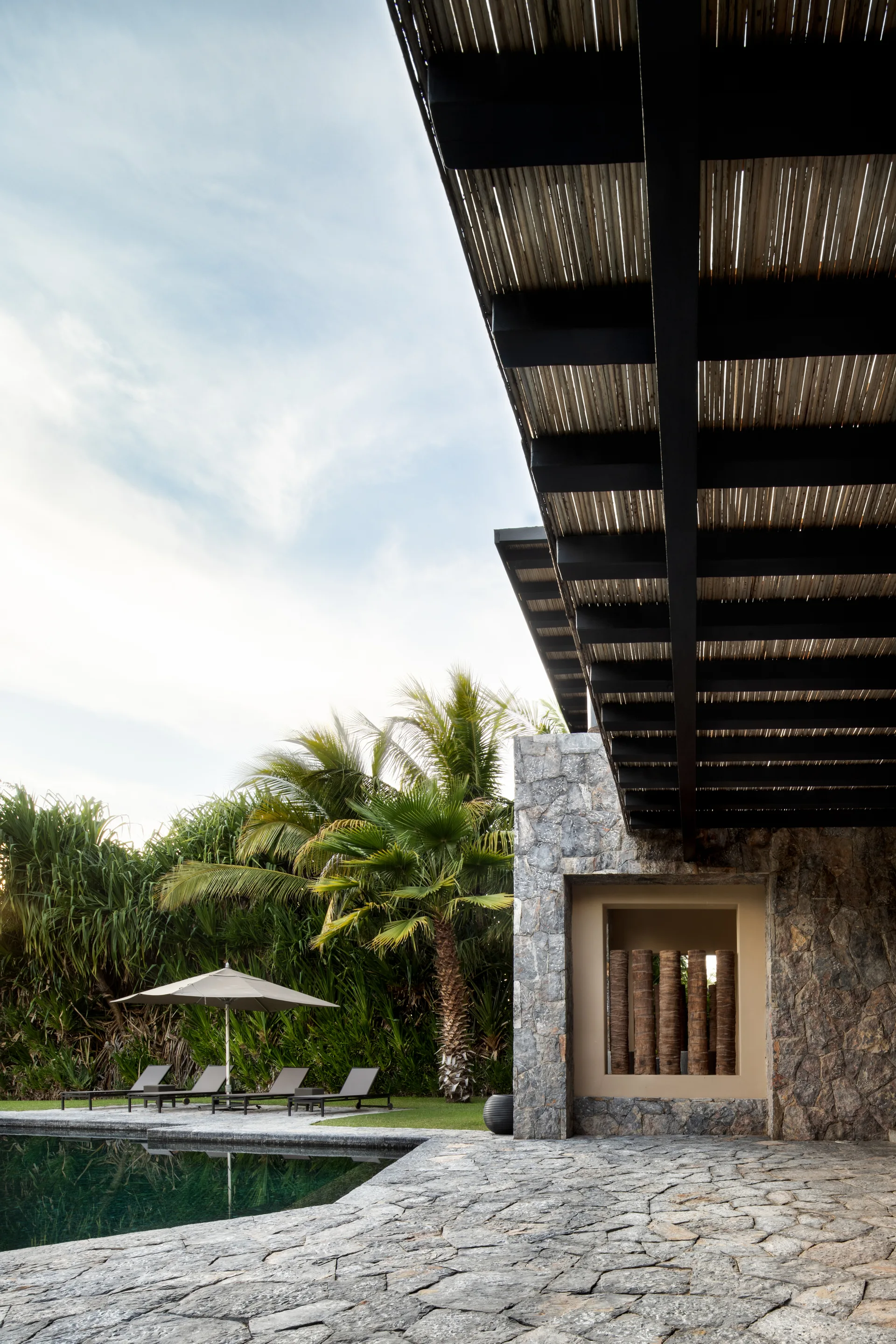 A tropical outdoor patio features an irregular stone floor, a stone wall with a recessed niche, and sun loungers next to a swimming pool.