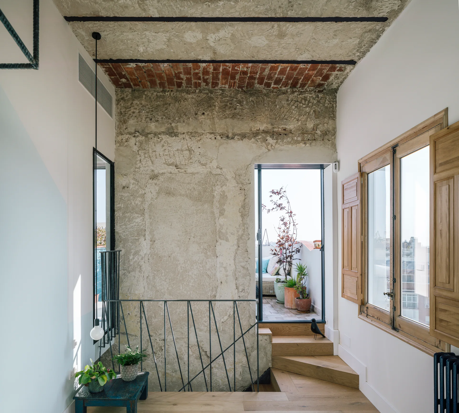 A wooden staircase with a dark metal railing leads to an outdoor terrace next to a raw concrete wall and an exposed brick ceiling.