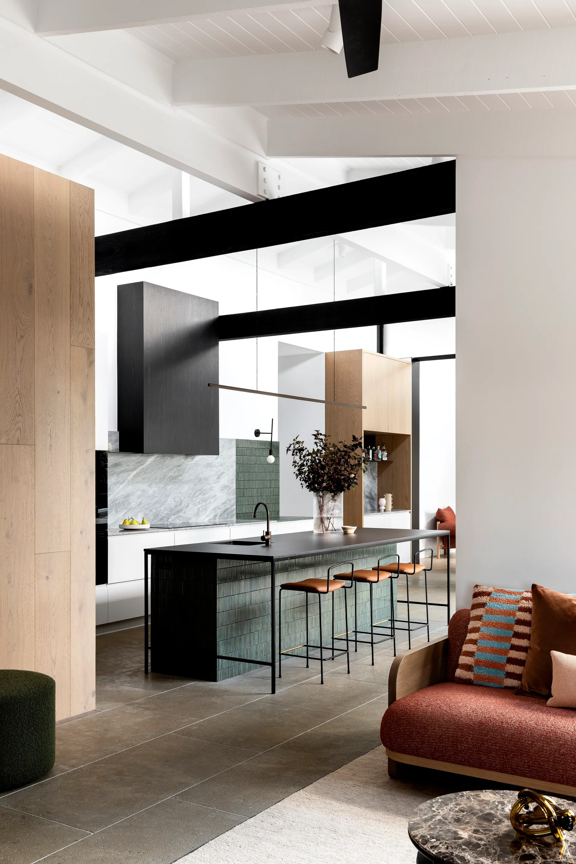 A contemporary open-plan kitchen features a green tiled island, black ceiling beams, wood cabinetry, and a terracotta sofa in the foreground.