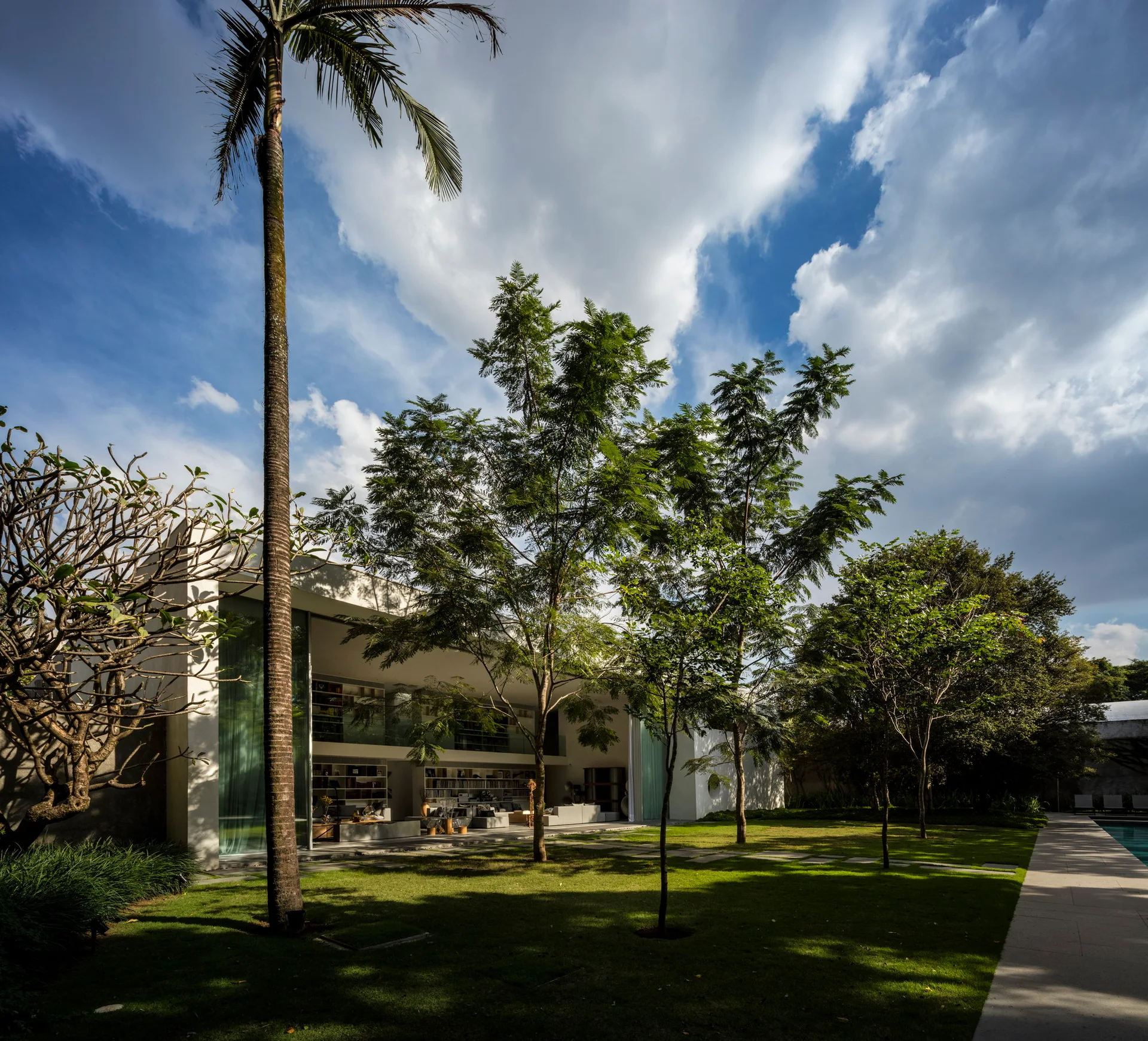 A modern white house with large glass walls stands behind a manicured green lawn with tall trees and a stone walkway next to a swimming pool.