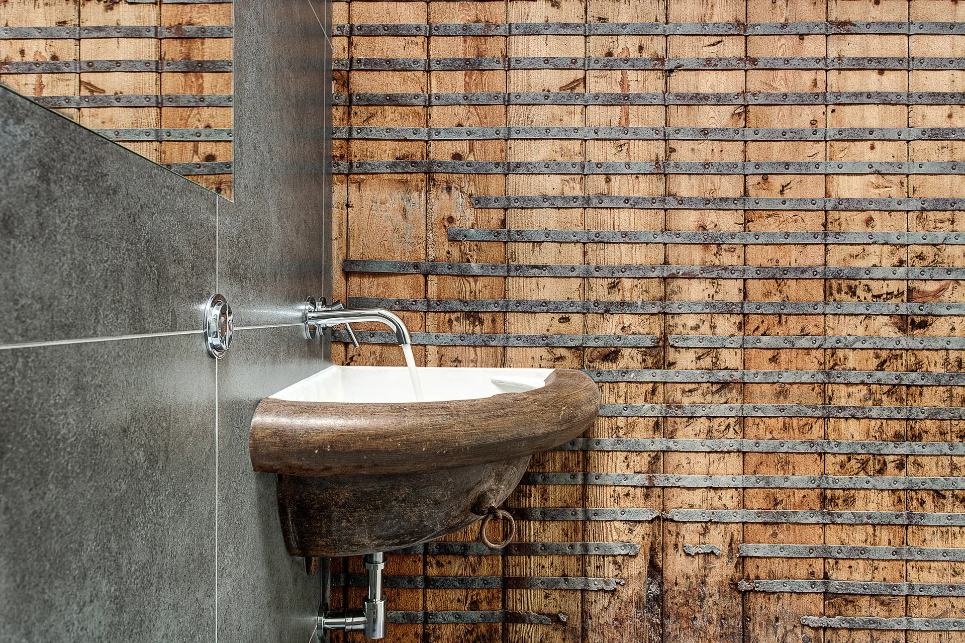 A rustic wall-mounted wooden sink with a chrome faucet is set against a background of dark grey tile and metal-banded wood planking.