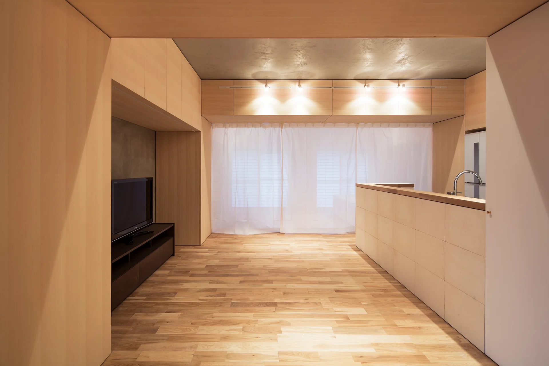 An open-plan room features light wood wall paneling, matching hardwood flooring, a tiled kitchen island, and a sheer white curtain.