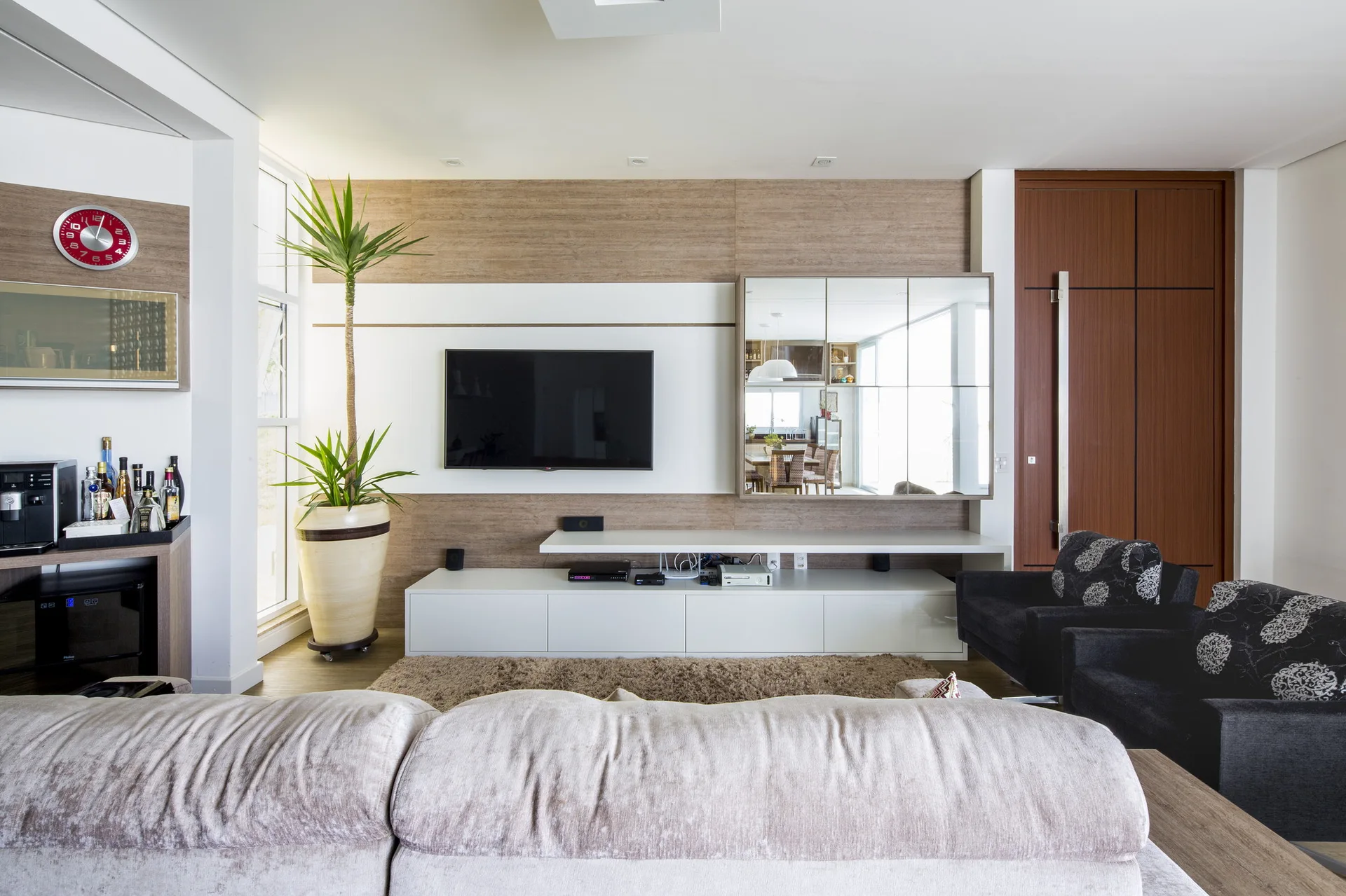 A modern living room features a flat-screen television mounted on a wood-paneled accent wall alongside a grid-patterned mirror and a low white media console.