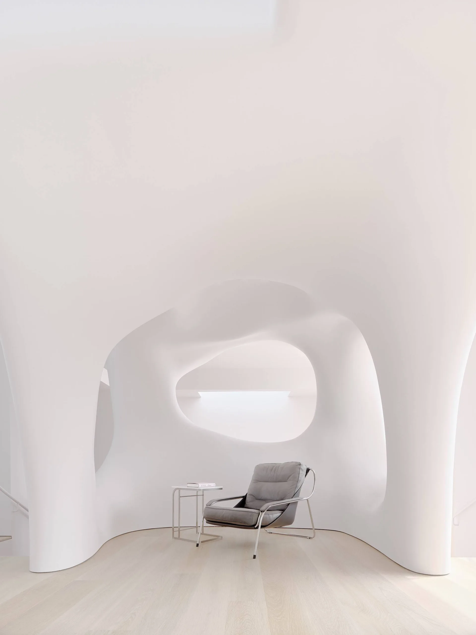 A grey minimalist armchair and small metal side table sit on light wood flooring inside a room with flowing, organic white sculptural plaster walls.