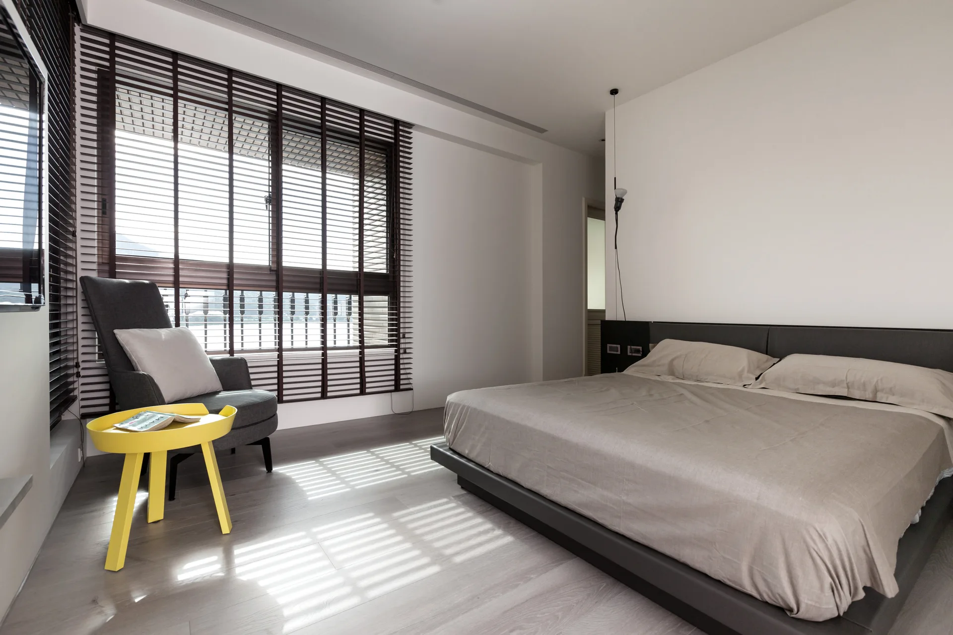 A minimalist bedroom features a grey low-profile bed, dark wood window blinds, a charcoal armchair, and a vibrant yellow tripod side table on light wood floors.