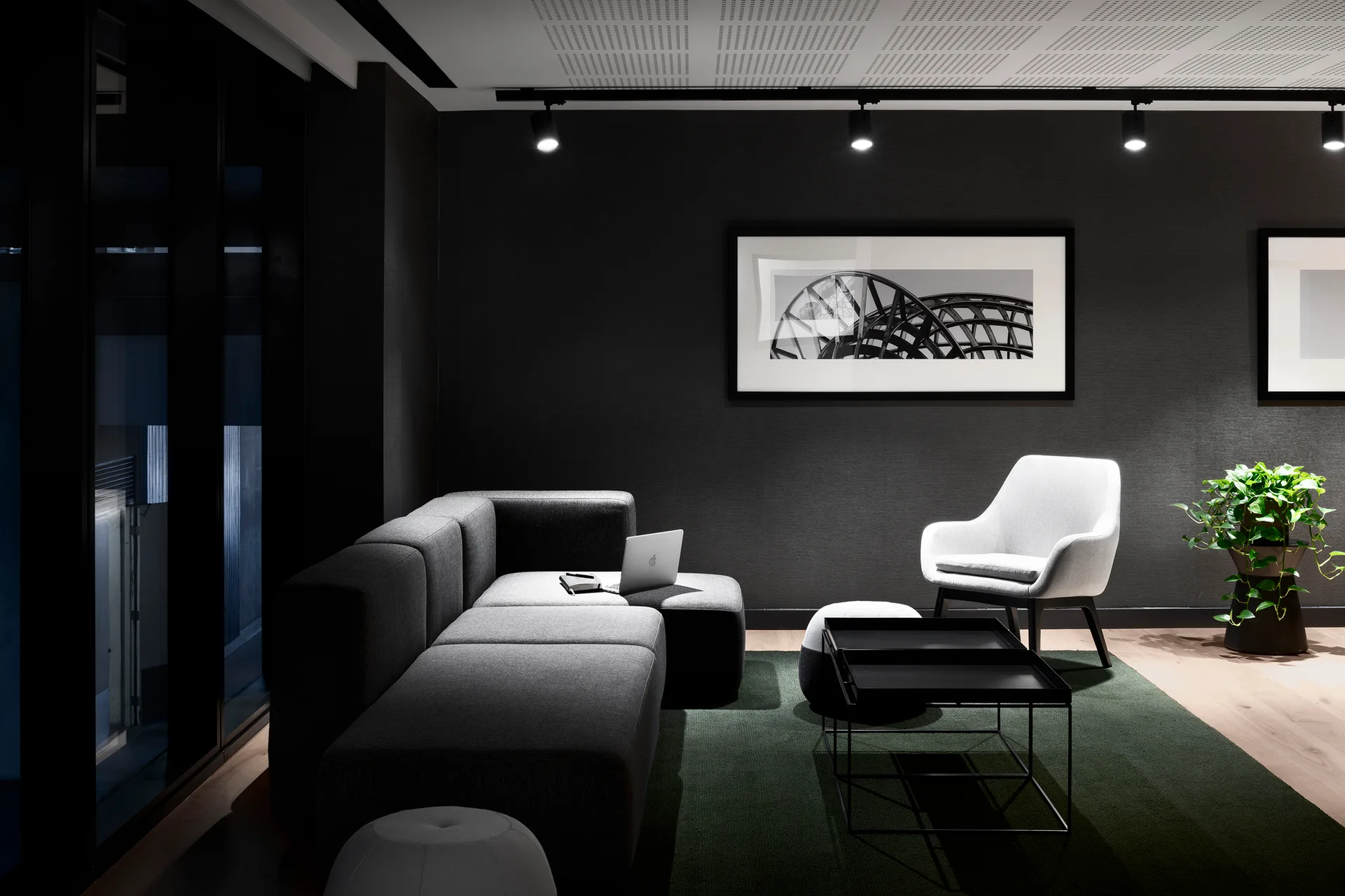 A dark gray minimalist lounge features a sectional sofa, a light accent chair, a green rug, and framed artwork illuminated by ceiling track lights.