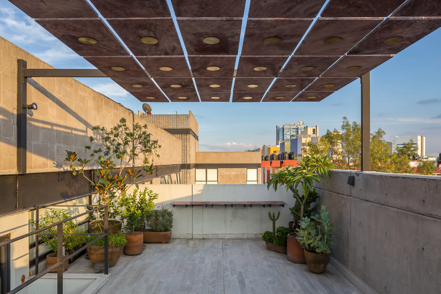 An outdoor rooftop terrace features a suspended grid canopy, concrete walls, grey stone flooring, and potted green plants in terracotta planters.