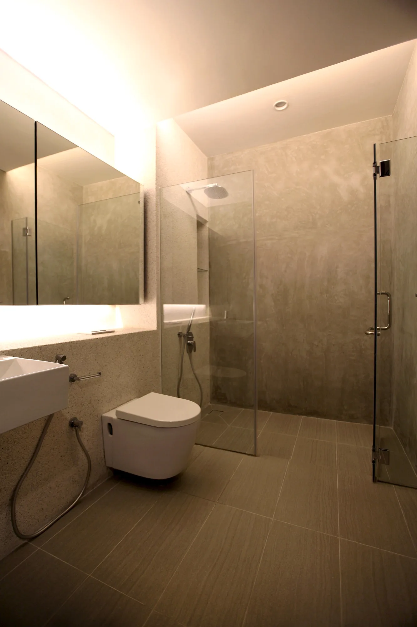 A modern bathroom features textured concrete walls, a wall-mounted toilet, a glass shower enclosure, and a frameless mirror with warm lighting.