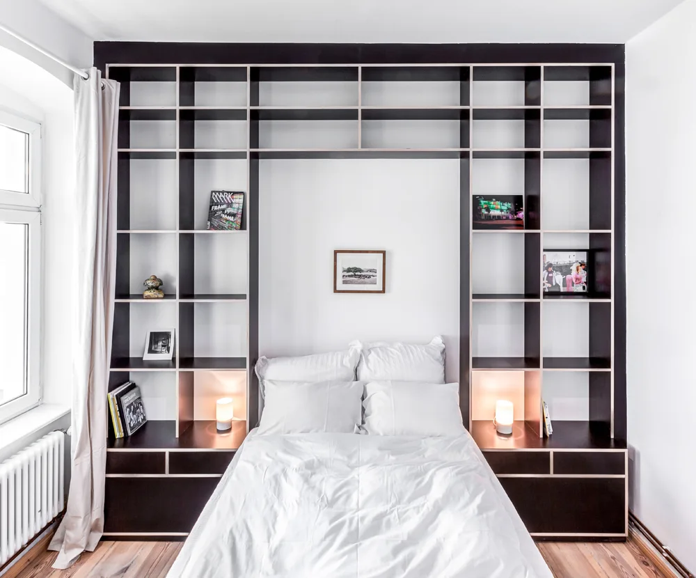 A white bed is recessed within a large black built-in storage unit featuring open shelves, plywood edges, and small illuminated lamps.