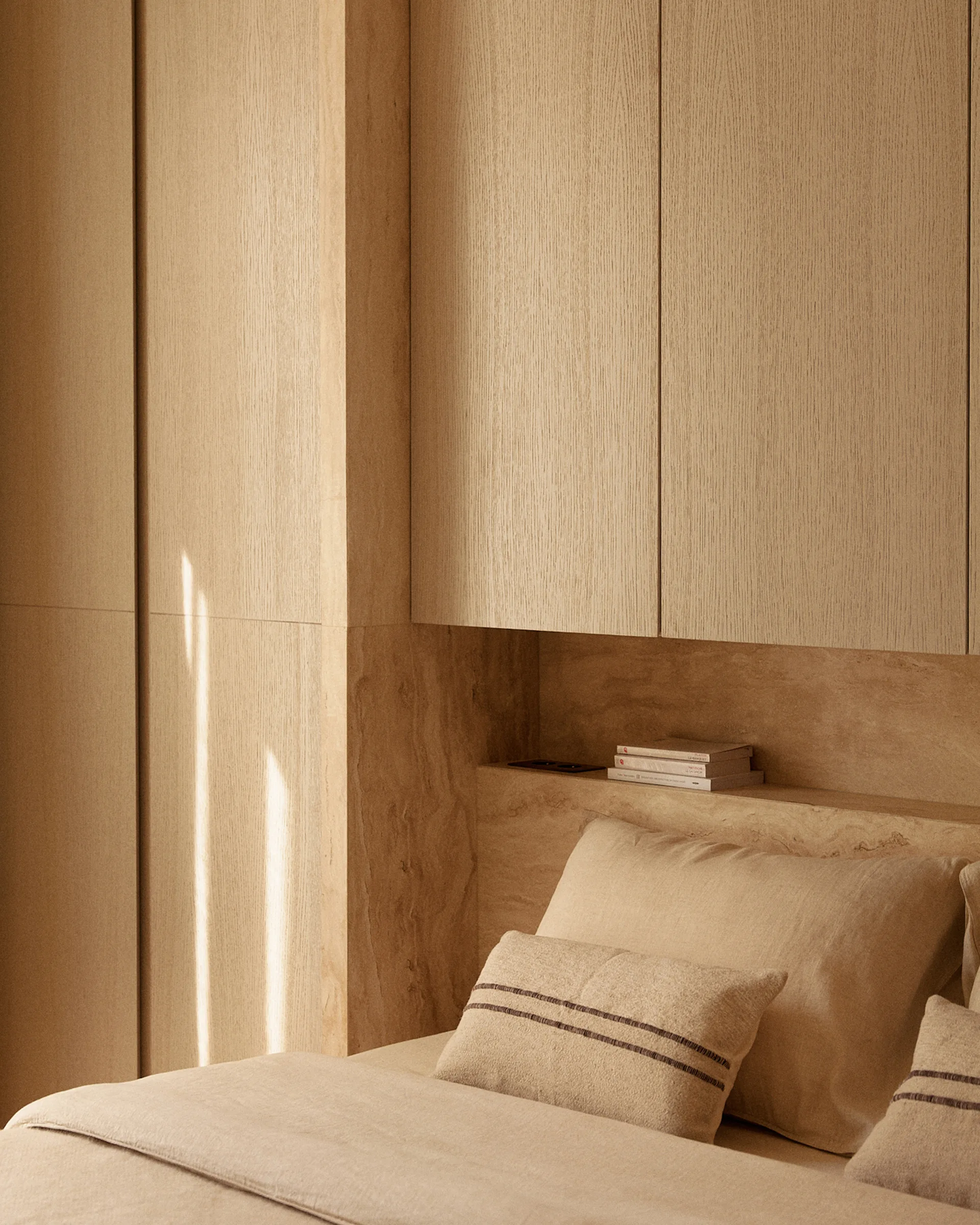 A minimalist bedroom featuring warm wood panel cabinets and a recessed stone alcove shelf above a bed with neutral linens.