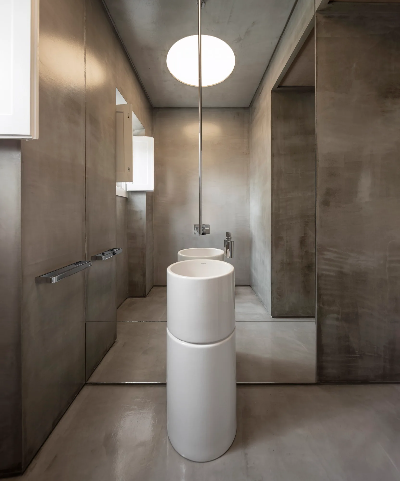 A minimalist grey cement bathroom featuring a white cylindrical pedestal sink, a mirrored wall, and a circular ceiling light.