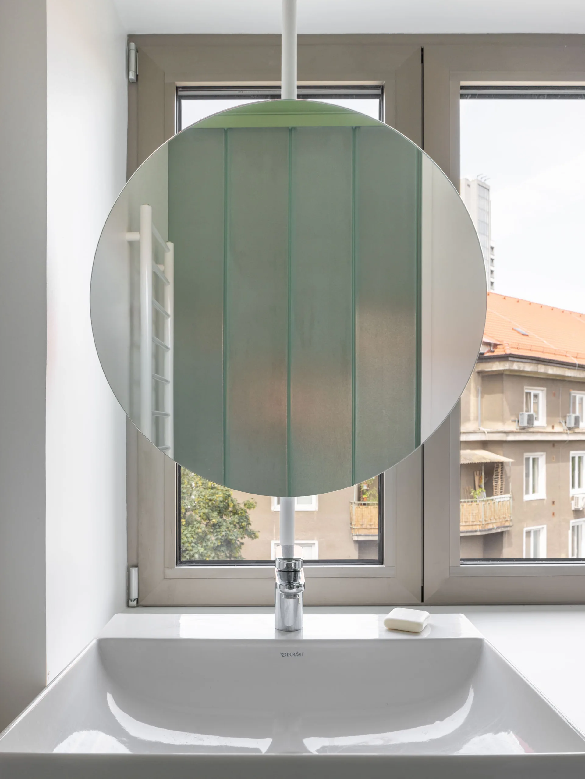 A circular mirror is mounted on a white vertical pole in front of a window, directly above a white ceramic bathroom sink with a chrome faucet.