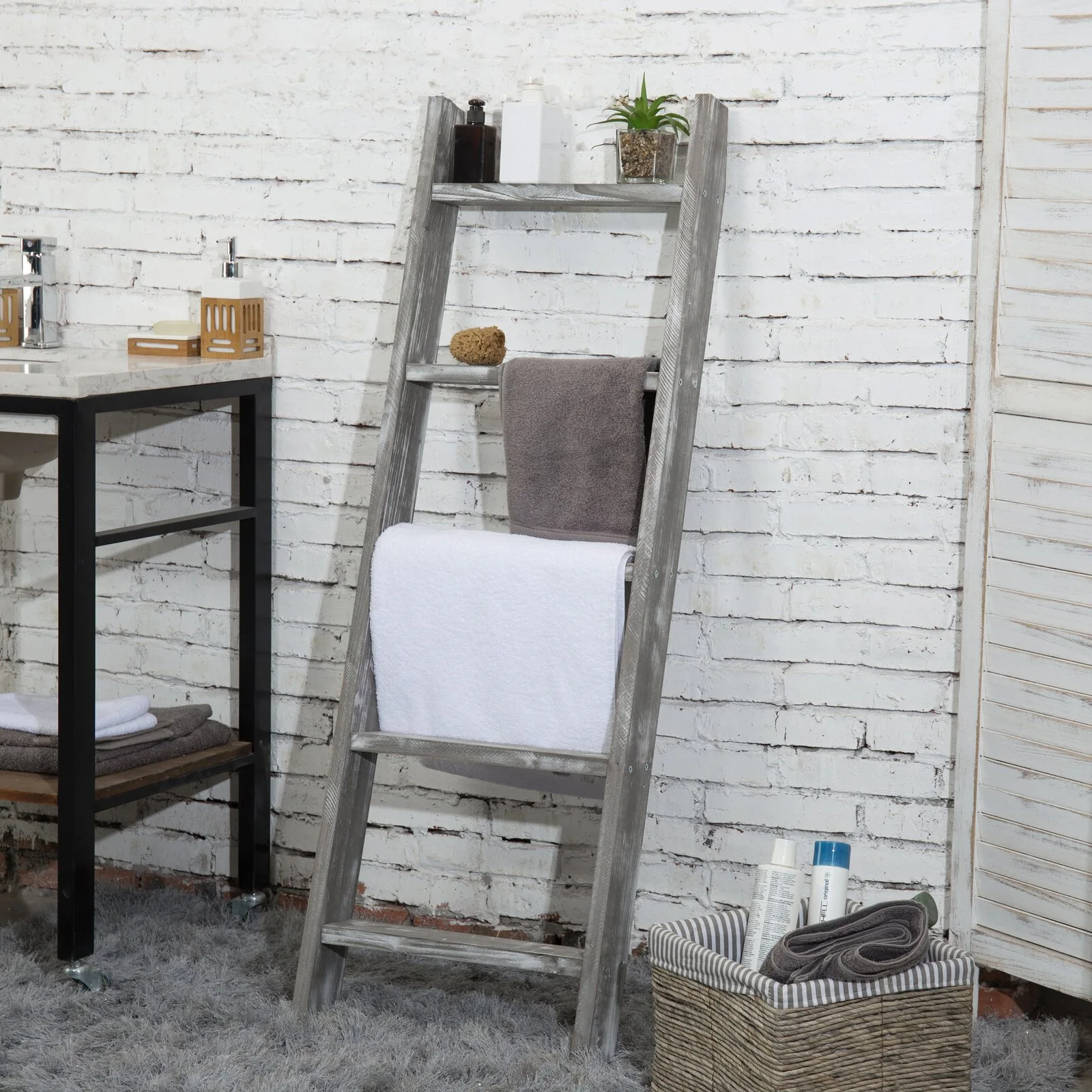 A grey weathered wooden leaning ladder shelf holding folded towels stands against a white painted brick wall next to a black-framed vanity.