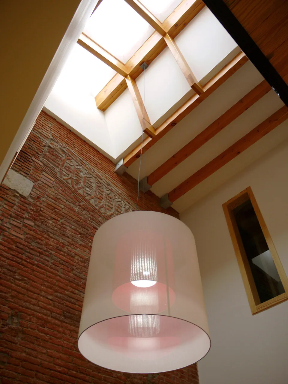 A large translucent cylindrical pendant light hangs in a tall room with an exposed red brick wall, a skylight, and wooden ceiling beams.