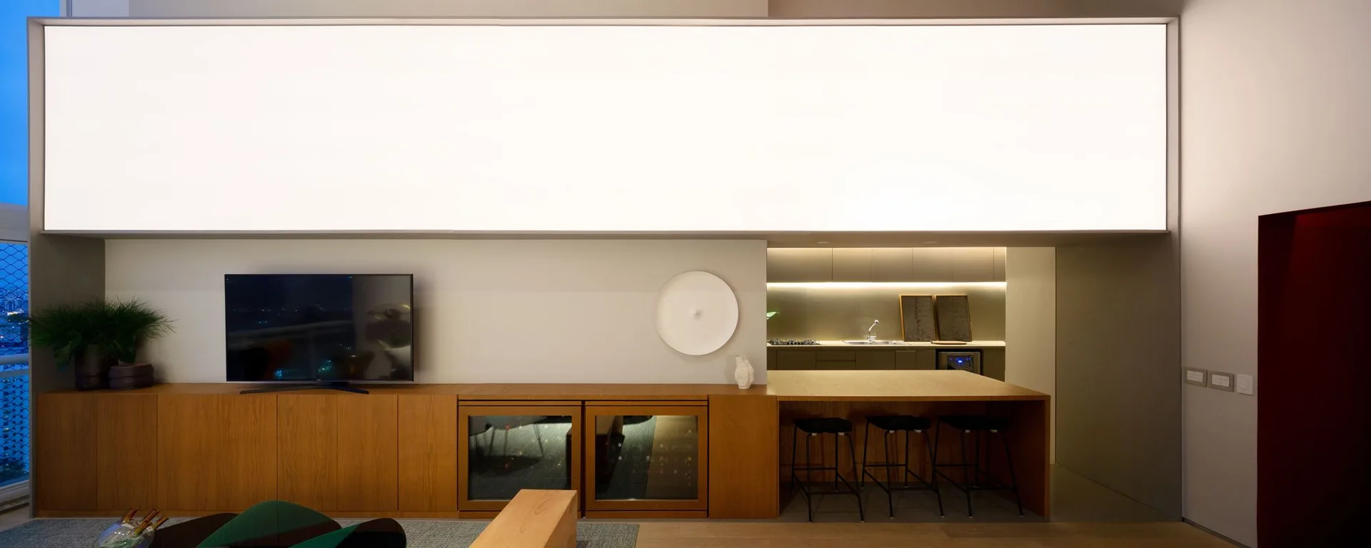 A long wood media console and matching bar counter sit beneath a massive horizontal glowing light panel in a minimalist interior.