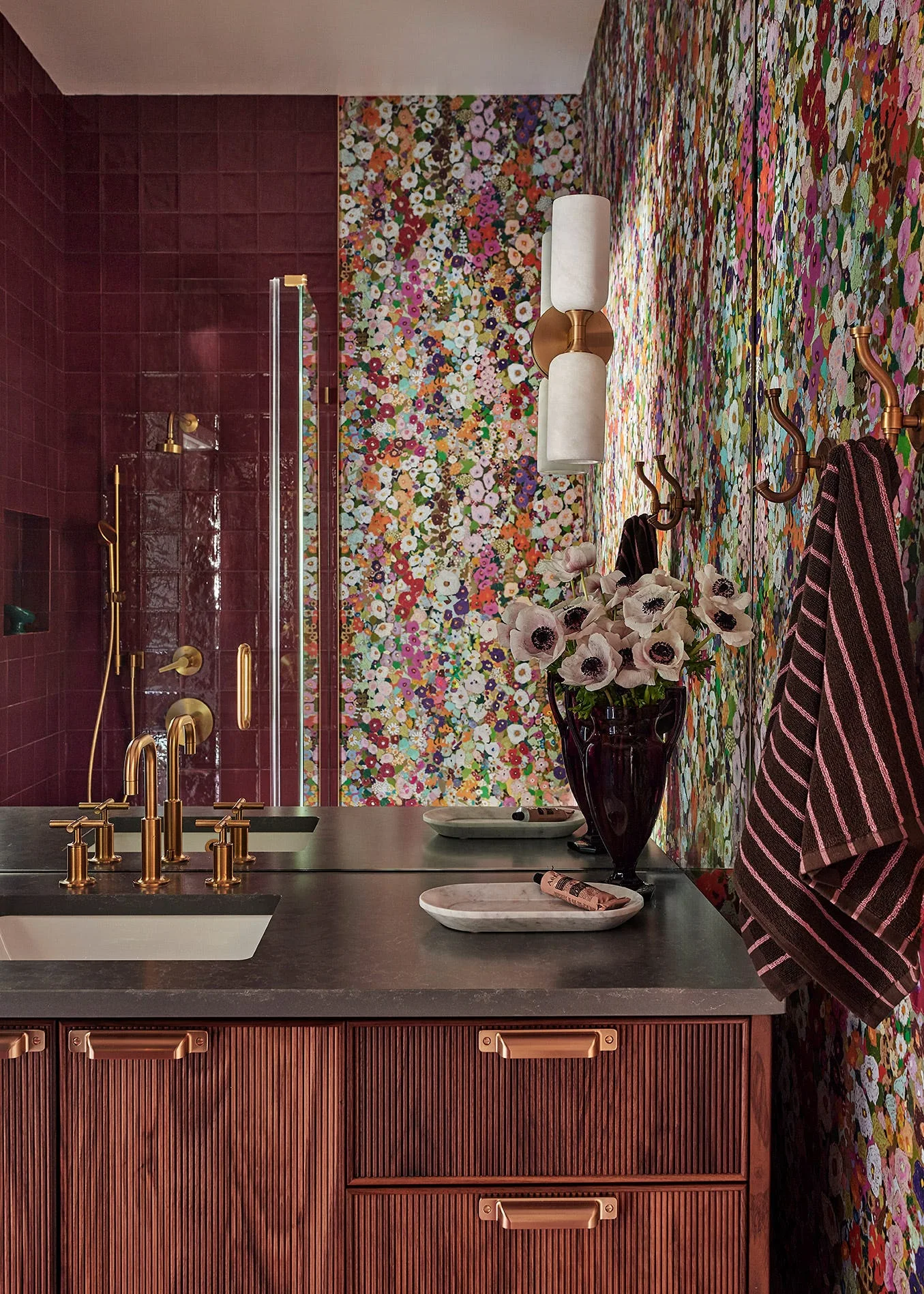 A bathroom vanity with a fluted wood cabinet and gold fixtures sits next to a shower with glossy maroon tiles and walls covered in floral wallpaper.
