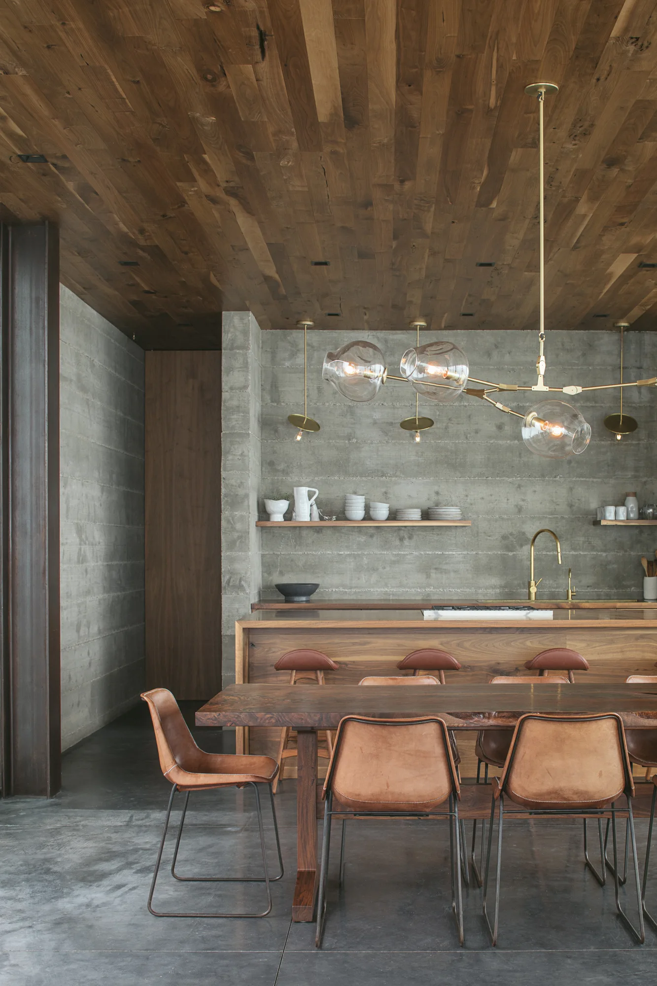 A modern kitchen and dining space features a wood plank ceiling, raw concrete walls, a wooden dining table with leather chairs, and brass light fixtures.