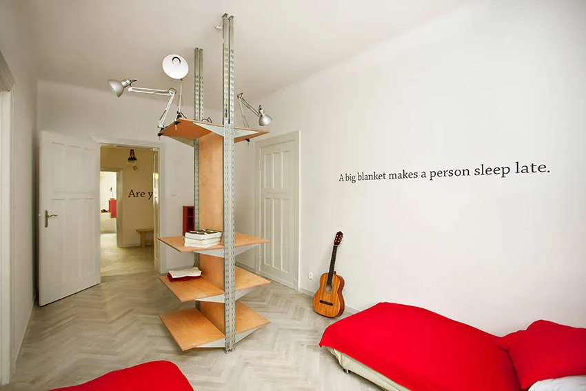 A minimalist white bedroom features a central floor-to-ceiling metal shelving unit, a bed with a red cover, and herringbone wood floors.