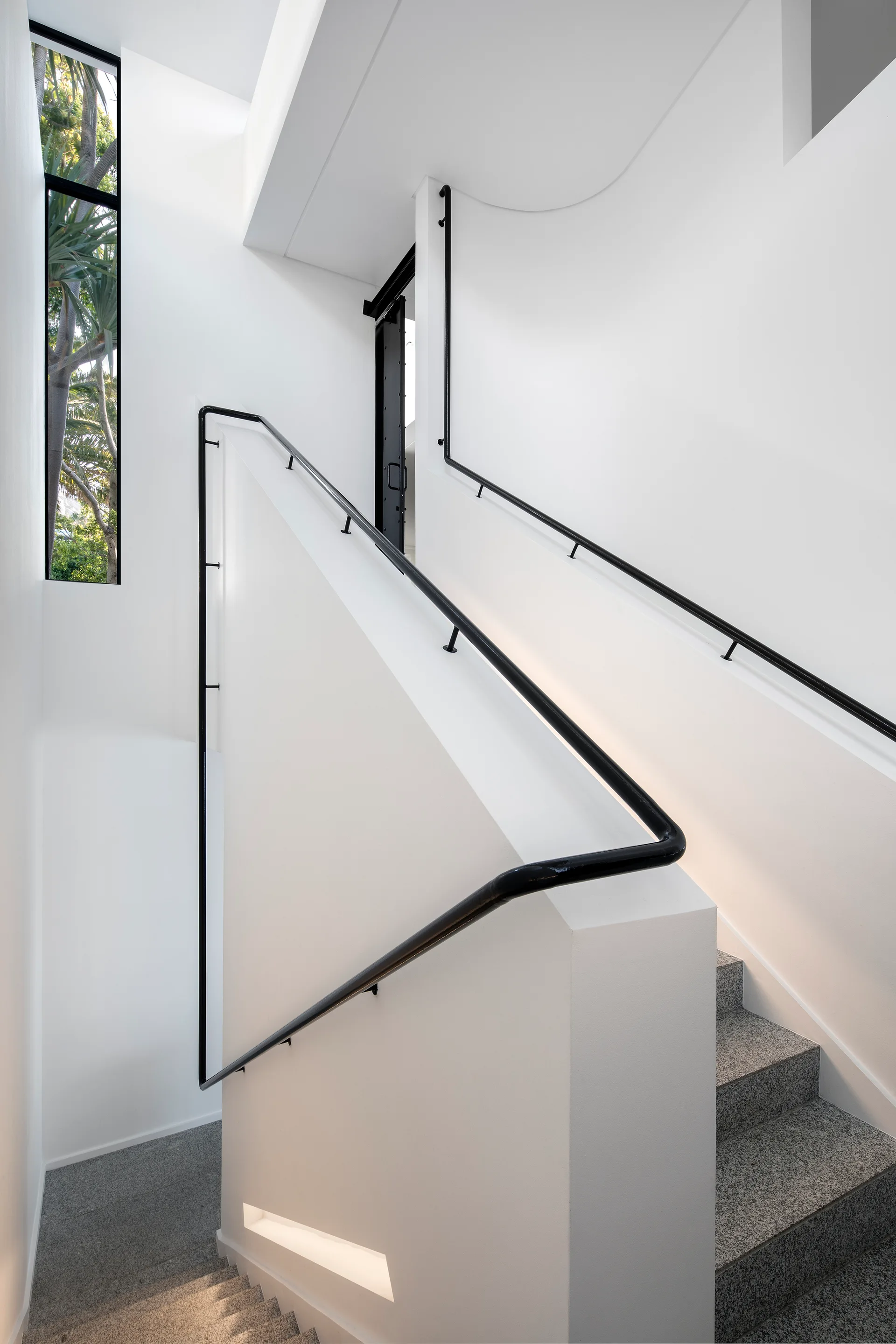 A minimalist white stairwell featuring grey speckled stone steps, a continuous black metal handrail, and a tall narrow window showing trees outside.