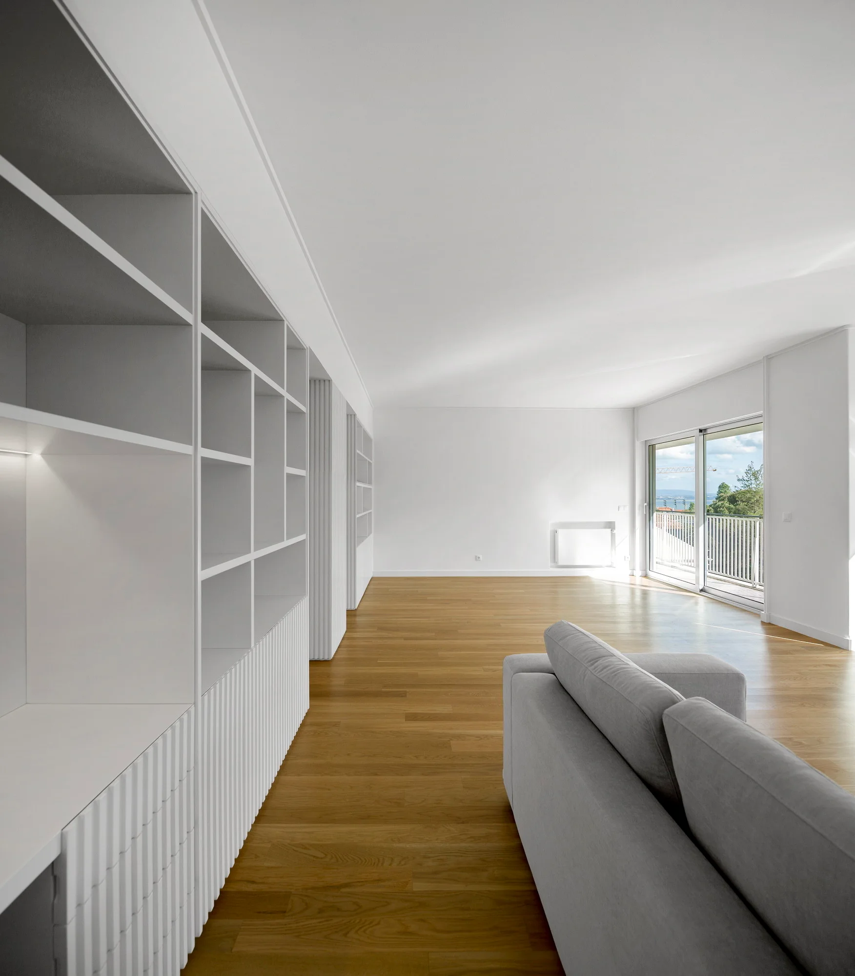 A minimalist living room features light hardwood floors, a grey sofa, and a large white built-in shelving unit with ribbed lower cabinet fronts.