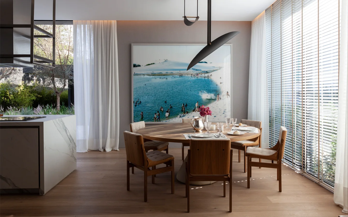 A round wooden dining table with chairs sits near a large window with blinds and a wall featuring a large framed beach photograph.