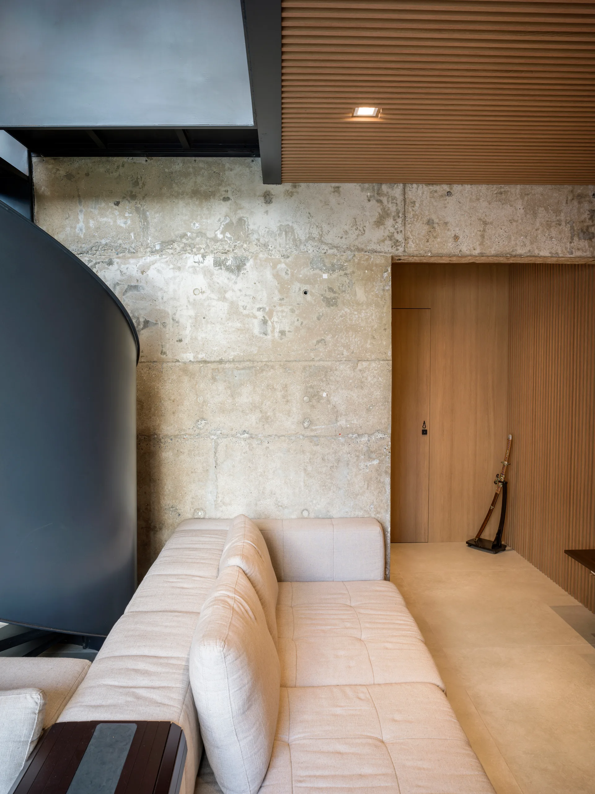 A cream modular sectional sofa sits in front of a raw concrete wall beneath a wood slatted ceiling and matching slatted wood panels.