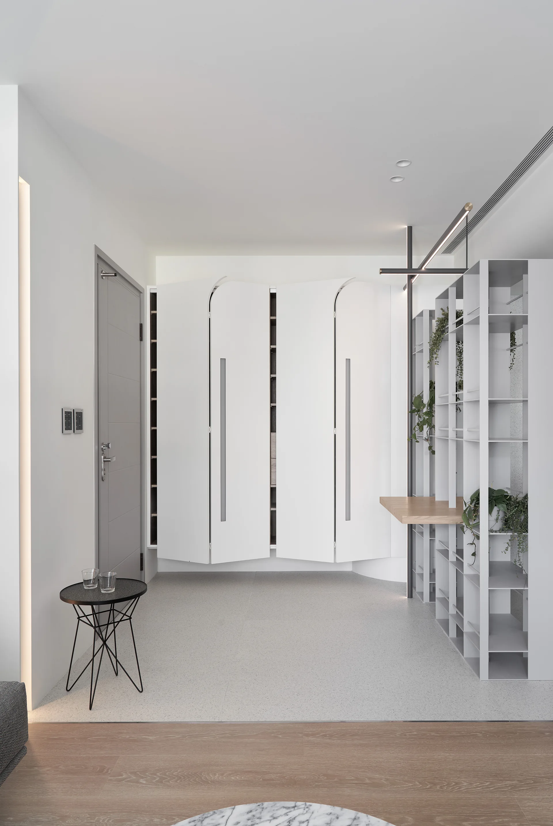 A minimalist white entryway featuring floating cabinets with curved doors, a tall metal shelving unit with an integrated wooden desk, and a grey door.