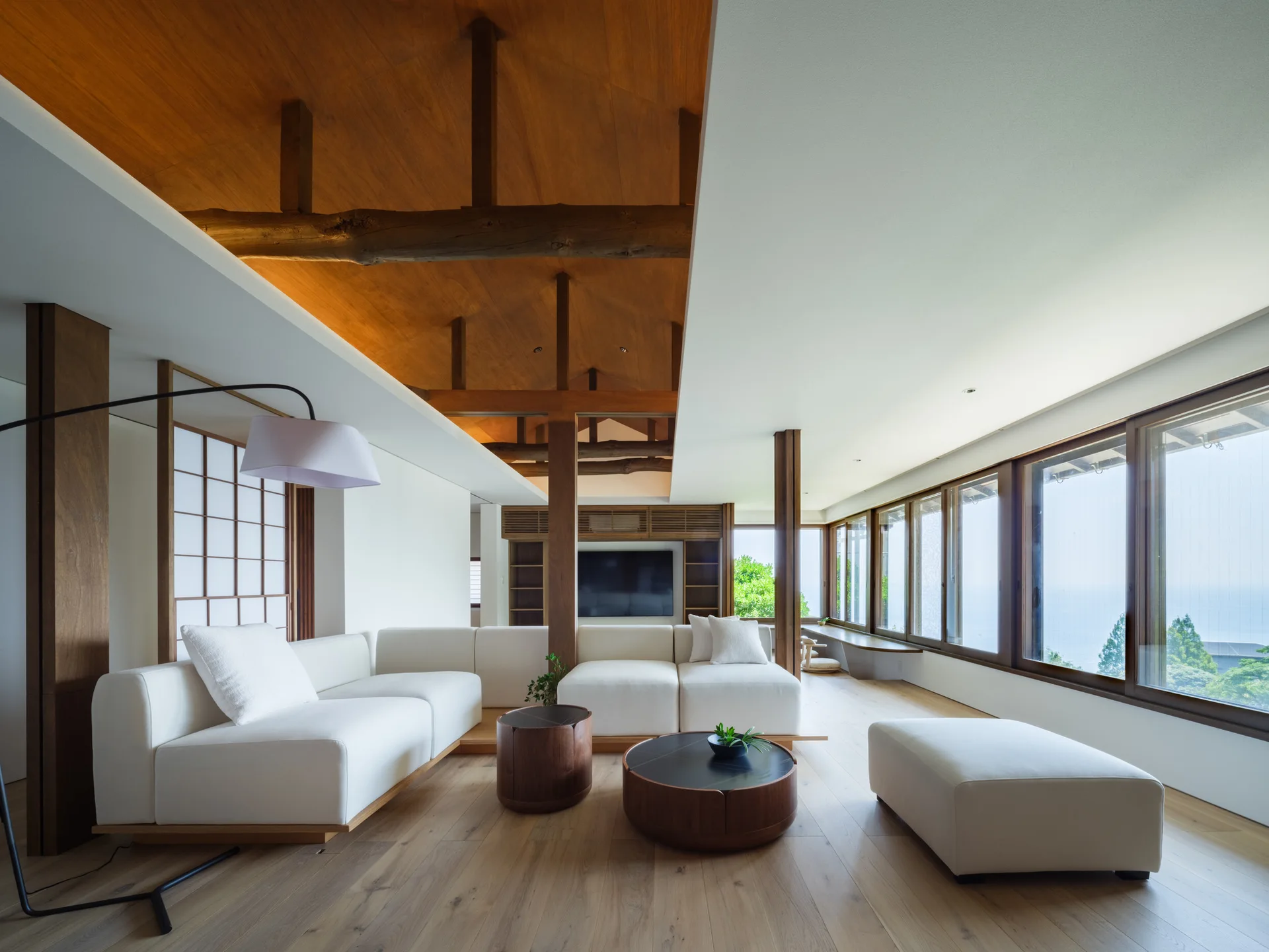 A minimalist living room featuring a white sectional sofa, round wood coffee tables, and a vaulted wood ceiling next to large windows overlooking the ocean.