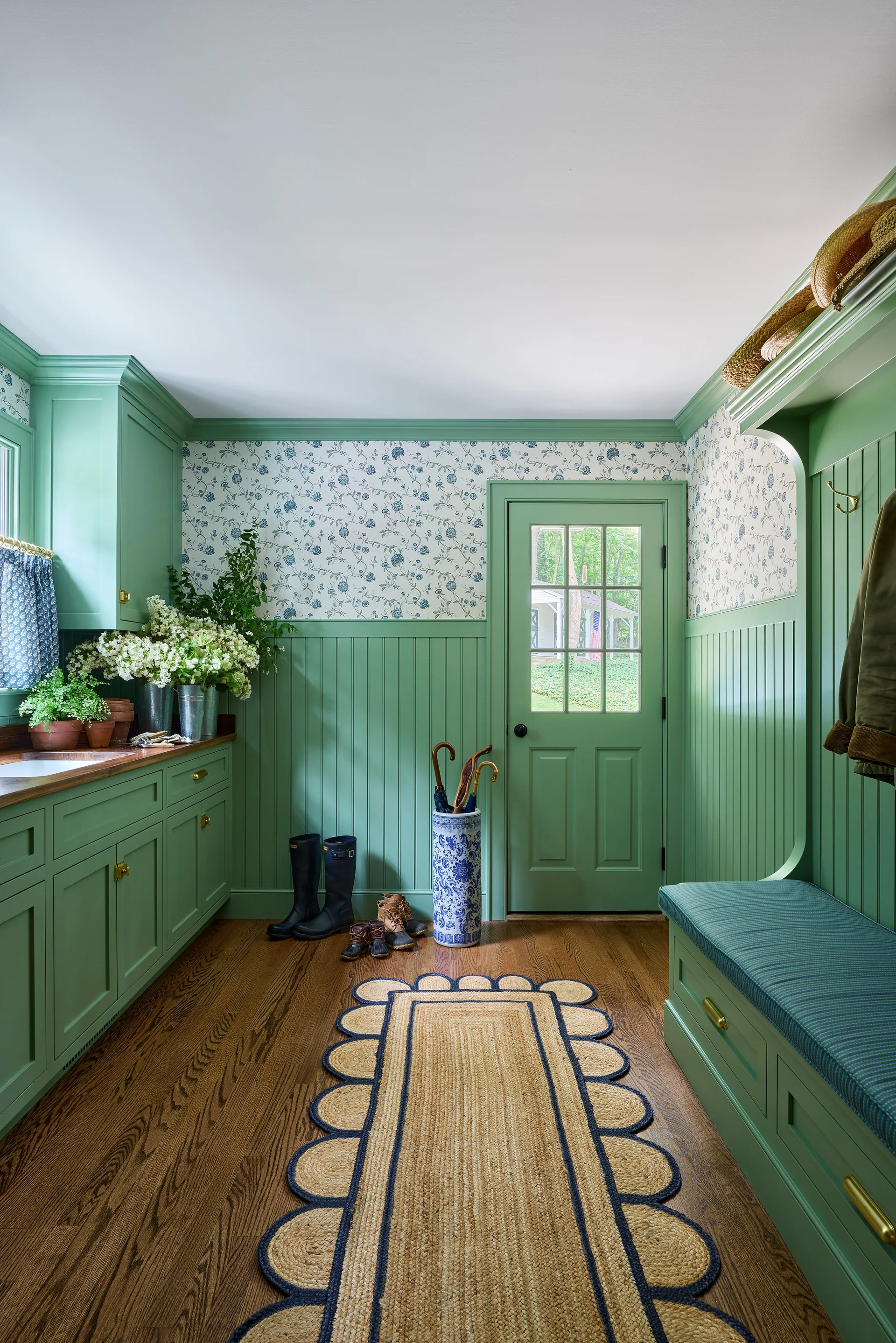 A mudroom featuring sage green beadboard walls, matching cabinetry, and floral wallpaper above a dark wood floor.