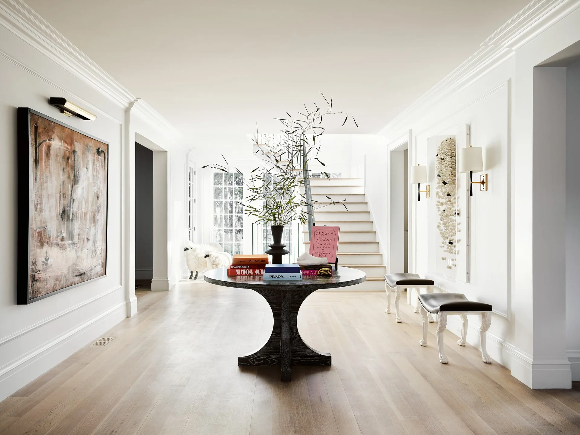A white paneled entryway with light wood floors features a central dark round table, a large abstract painting, and a staircase in the back.