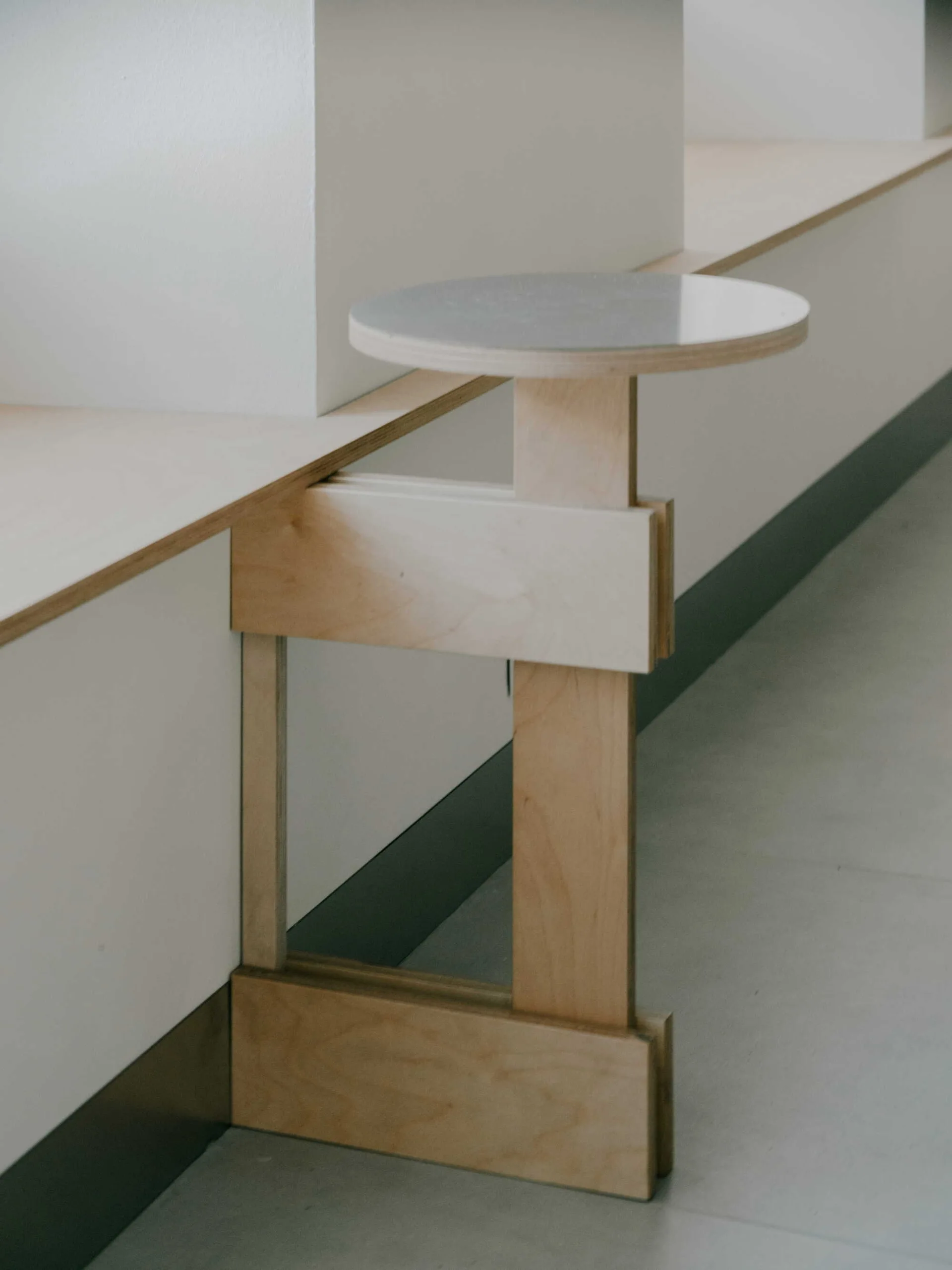 A minimalist circular wooden stool with a geometric base integrated into a long white built-in bench.