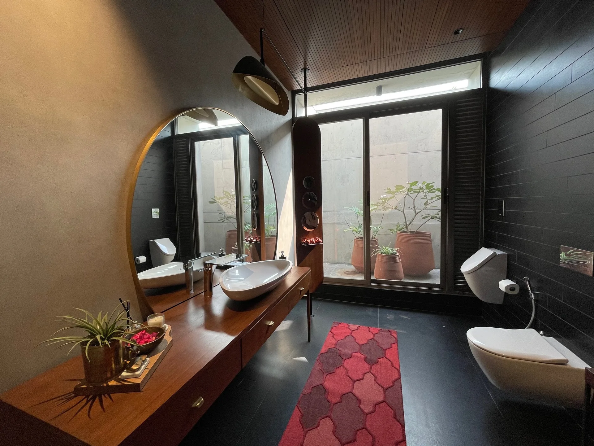 A modern bathroom features a long wood vanity, a circular mirror, a white vessel sink, black tiled walls, and a large window facing outdoor potted plants.