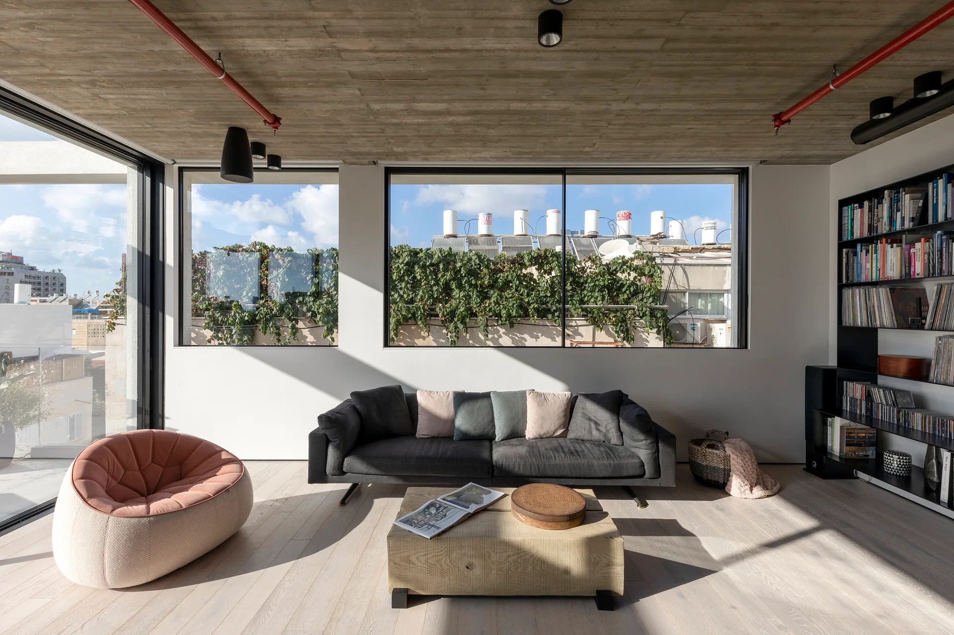 A sunlit living room features a gray sofa, a pink round armchair, a low wood coffee table, and an exposed concrete ceiling with red pipes.