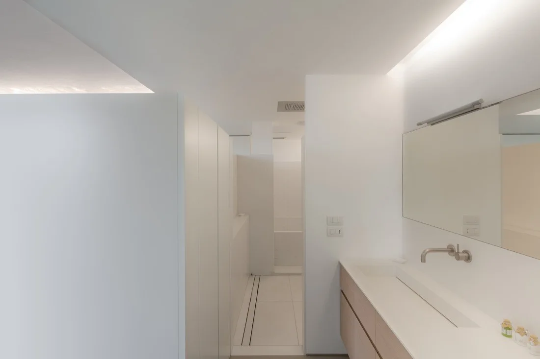 A minimalist white bathroom featuring a light wood vanity with a wall-mounted faucet, a frameless mirror, and integrated cove lighting.