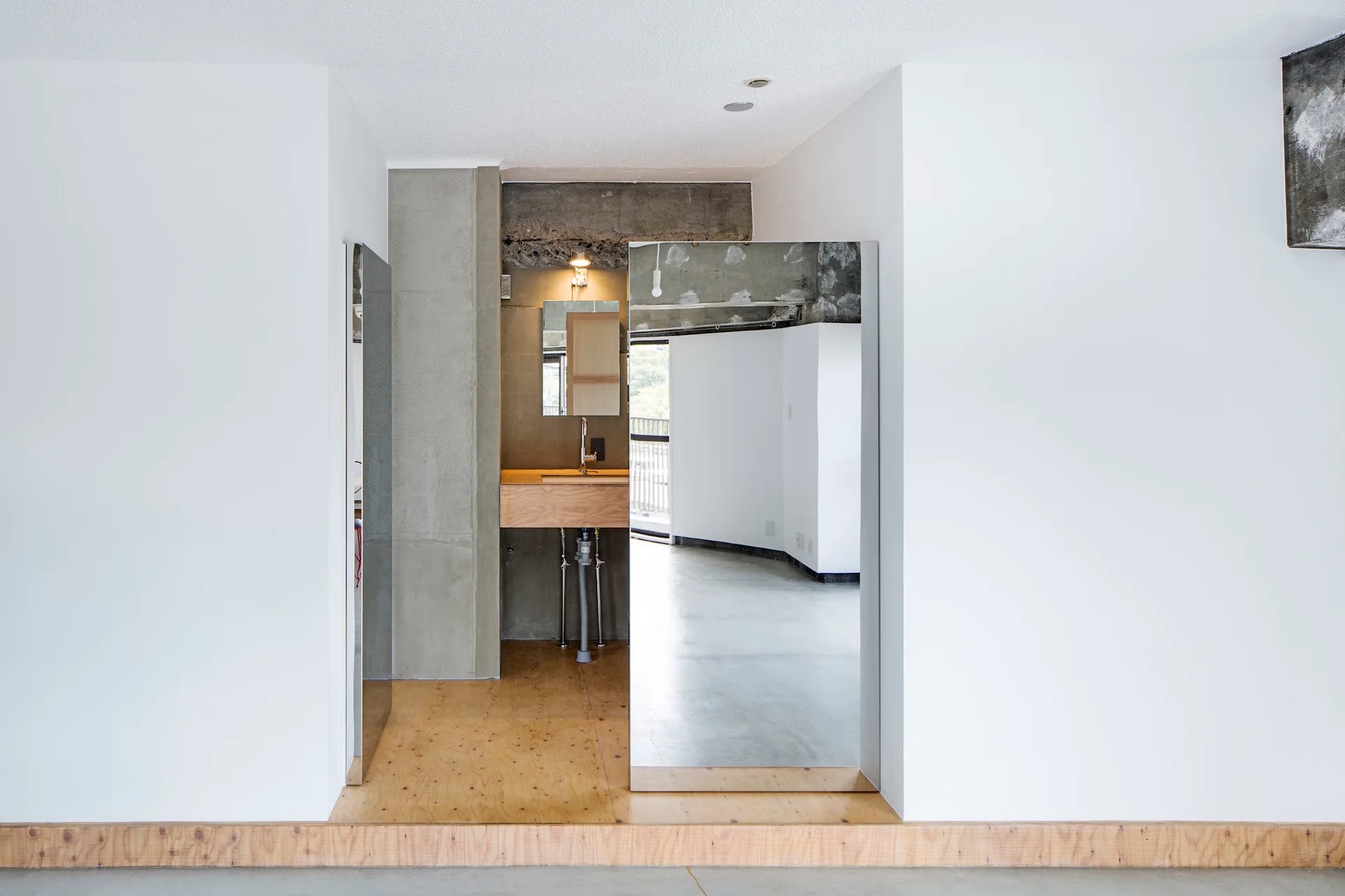 An industrial-style bathroom alcove featuring a light wood vanity, raw concrete walls, exposed plumbing, and chrome fixtures.