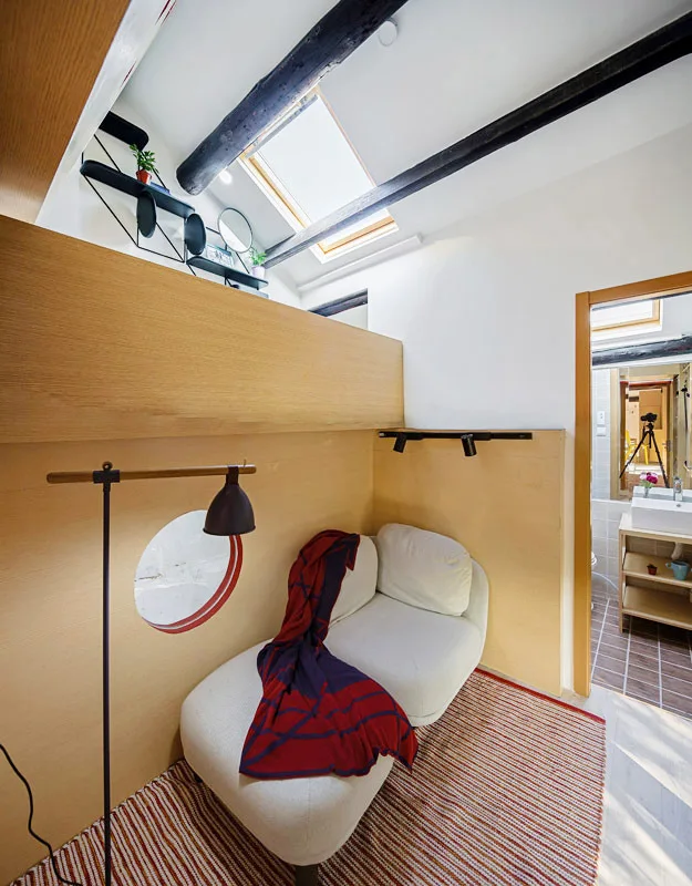 A white chaise lounge sits in an attic reading nook next to a wood-paneled partition wall with a round cutout beneath a sloped ceiling skylight.