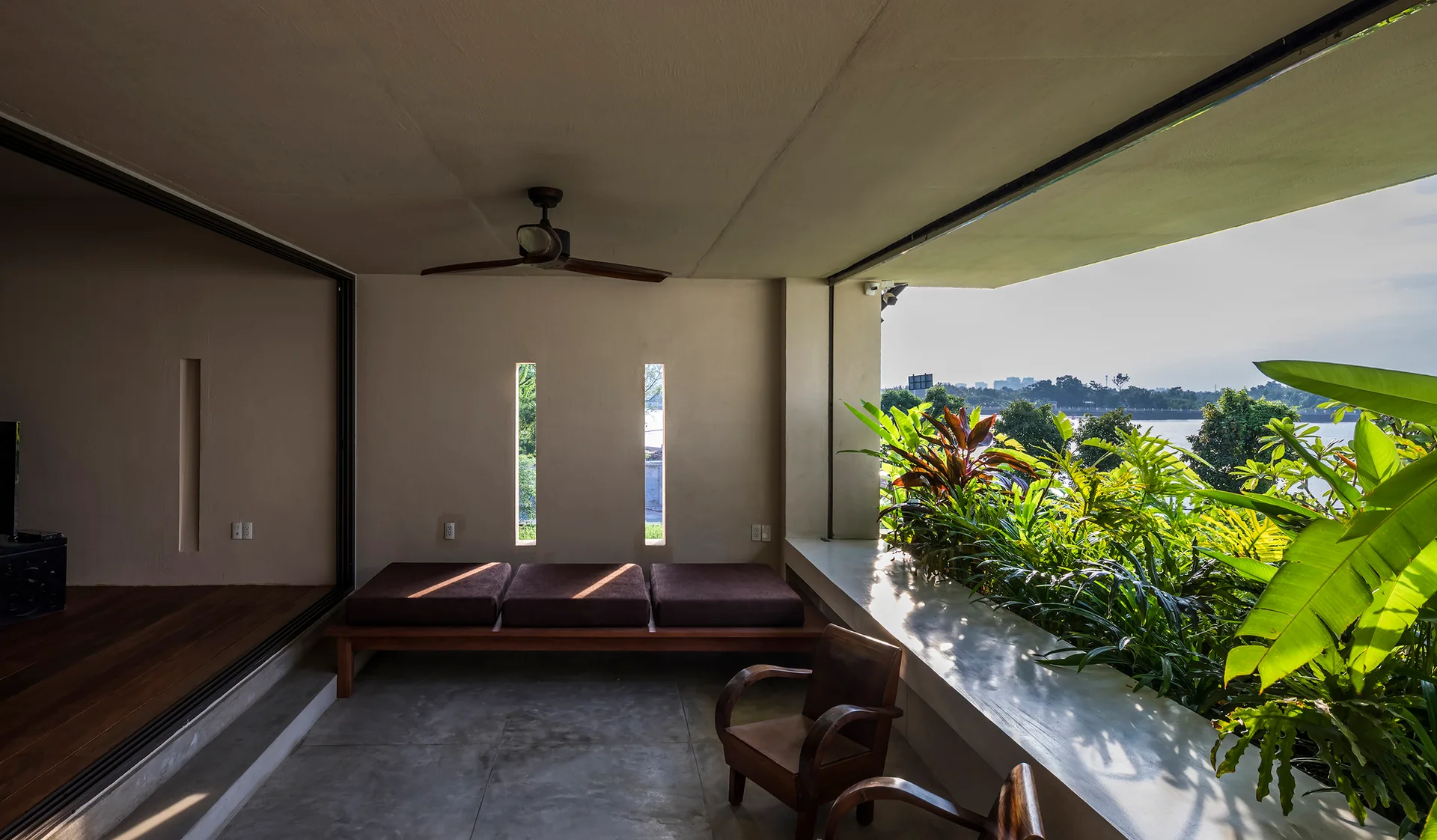 A covered concrete patio featuring a wooden daybed, wooden armchairs, and a built-in white planter ledge overflowing with tropical green foliage.