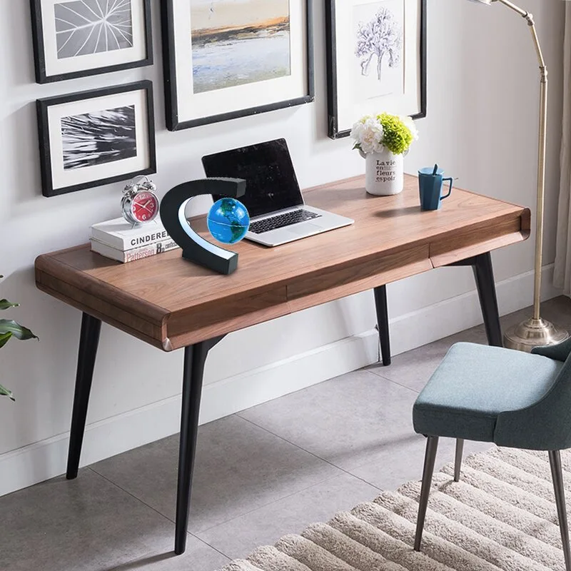 A wooden writing desk with a laptop stands against a white wall decorated with framed art, paired with an upholstered blue chair on a textured rug.