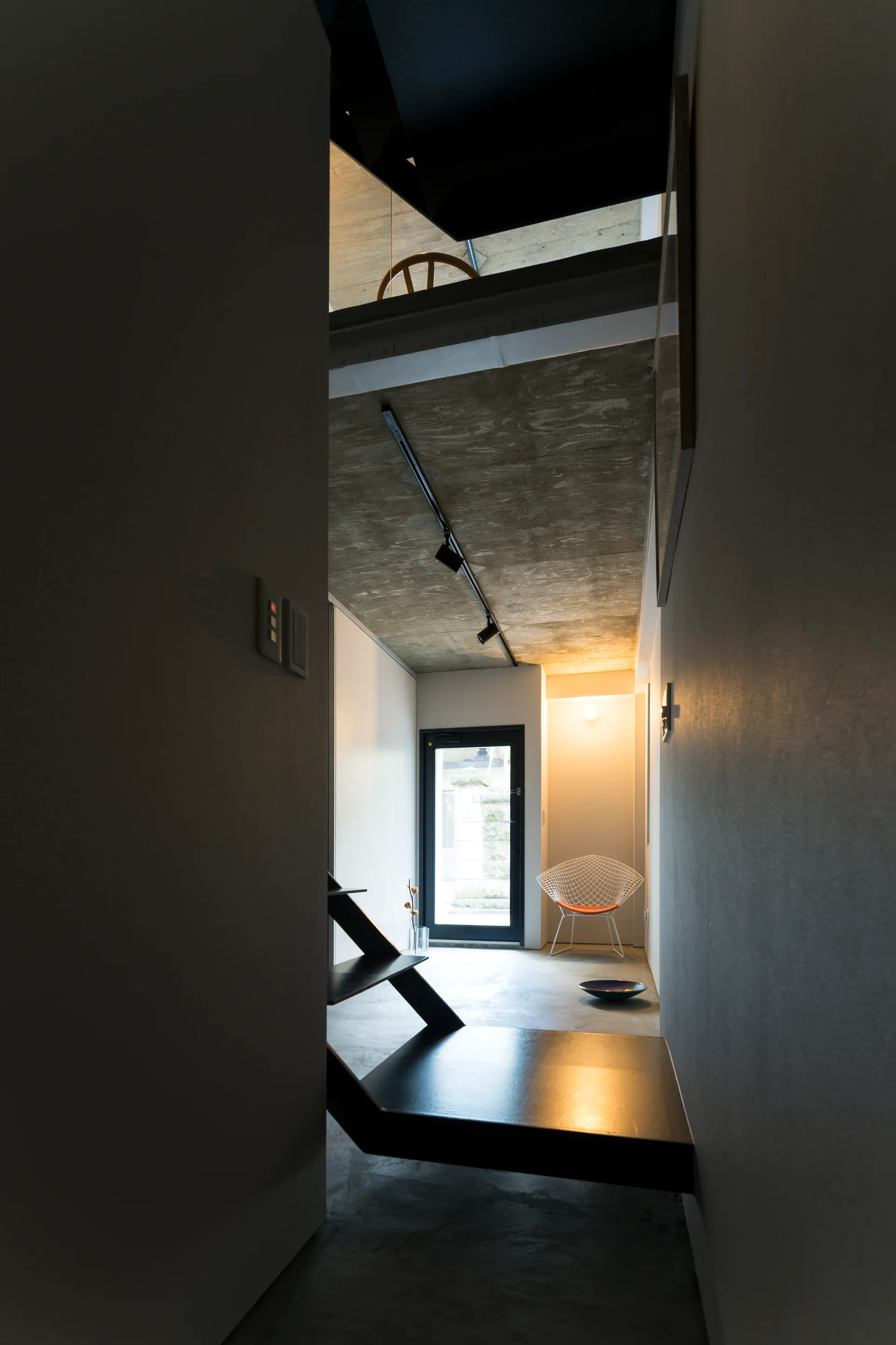 A minimalist entryway with an angular black metal staircase, a raw concrete ceiling, and a white wire chair near a glass door.