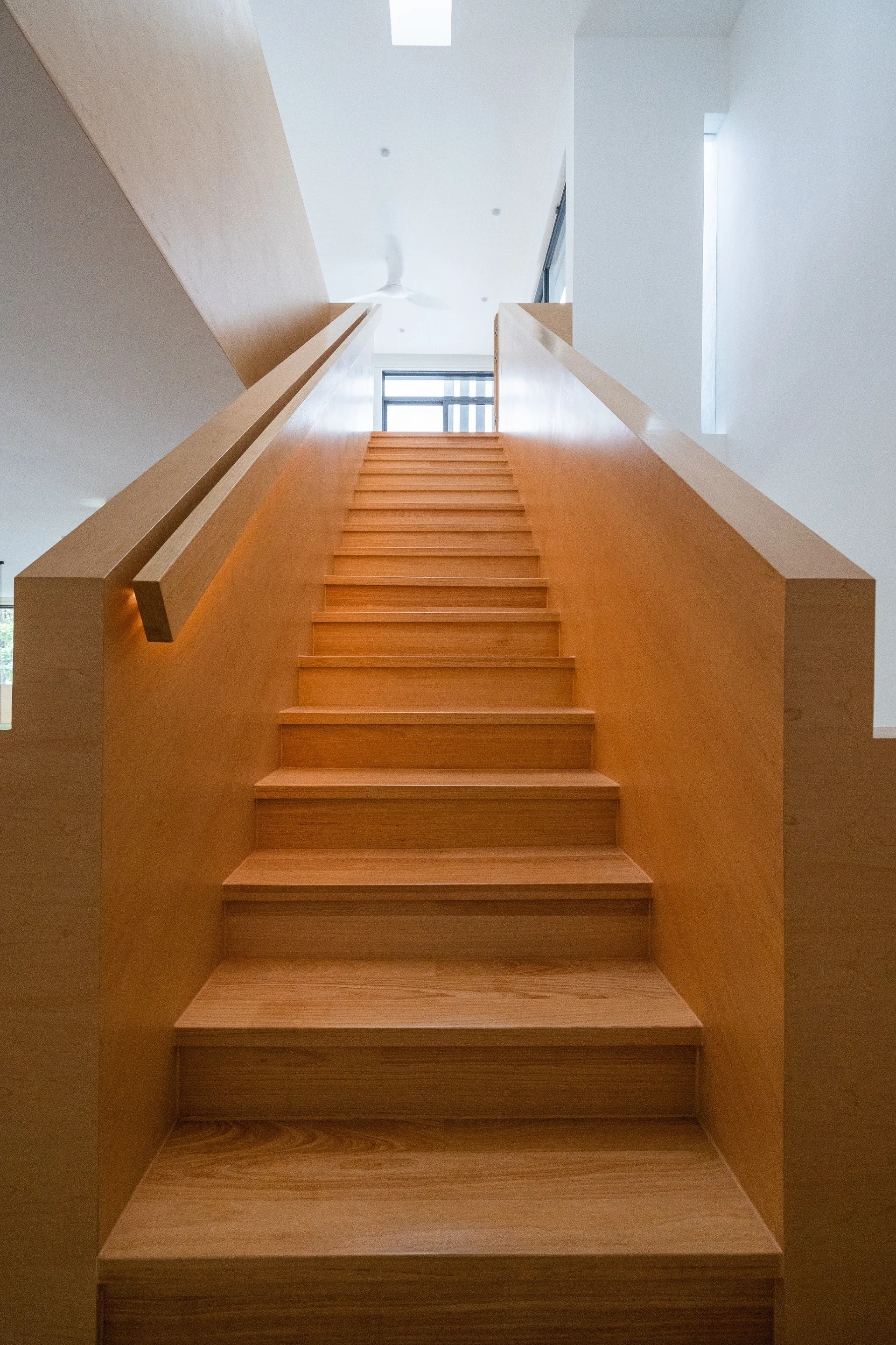An upward view of a minimalist wooden staircase flanked by solid wood handrails under a bright white ceiling.