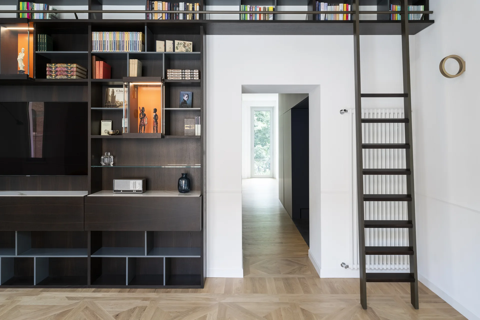 A dark wood built-in shelving unit with a rolling library ladder wraps around a central doorway over light wood parquet flooring.