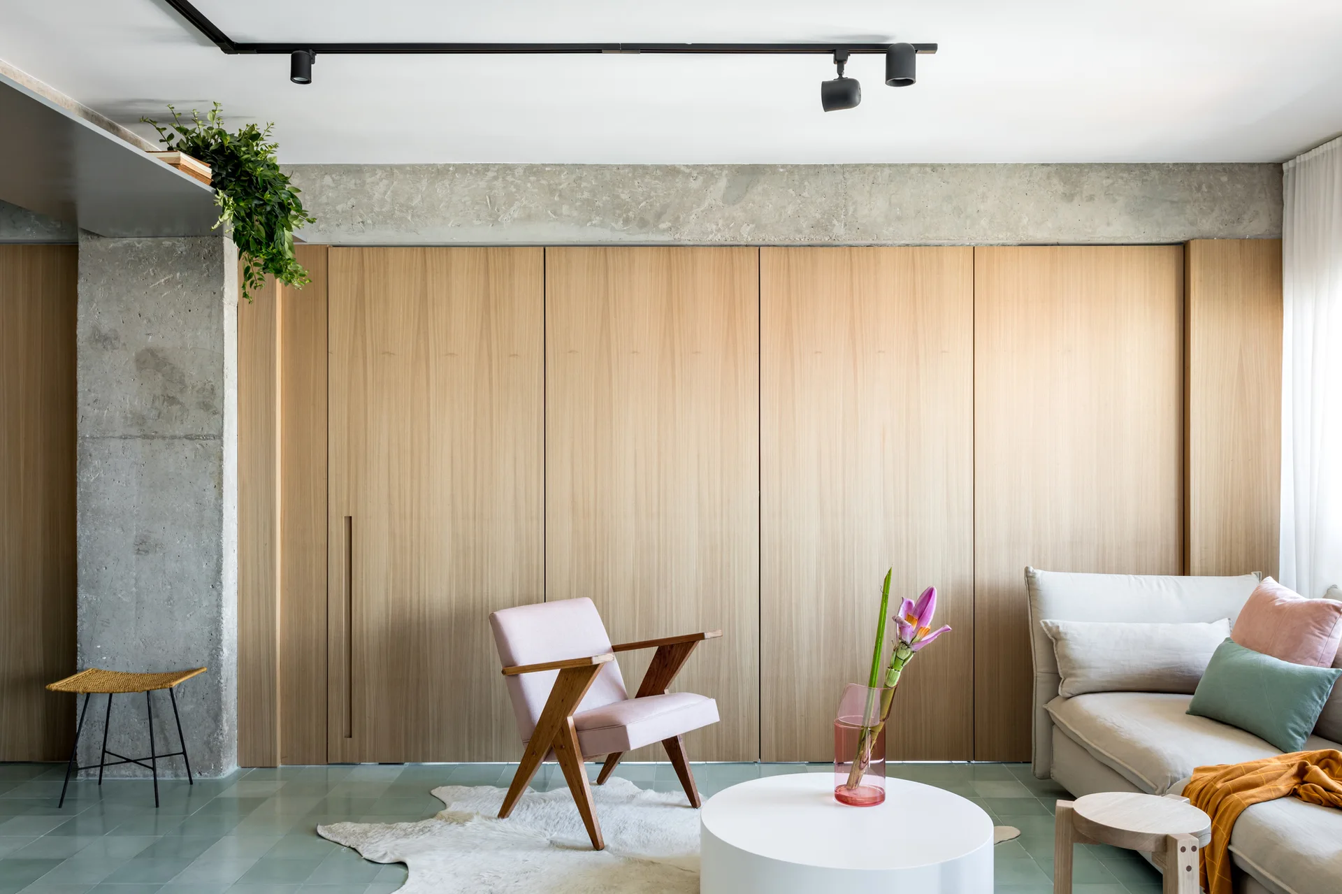 A modern living room features a full-height light wood paneled wall, an exposed concrete beam, a pink armchair, and a mint green tiled floor.